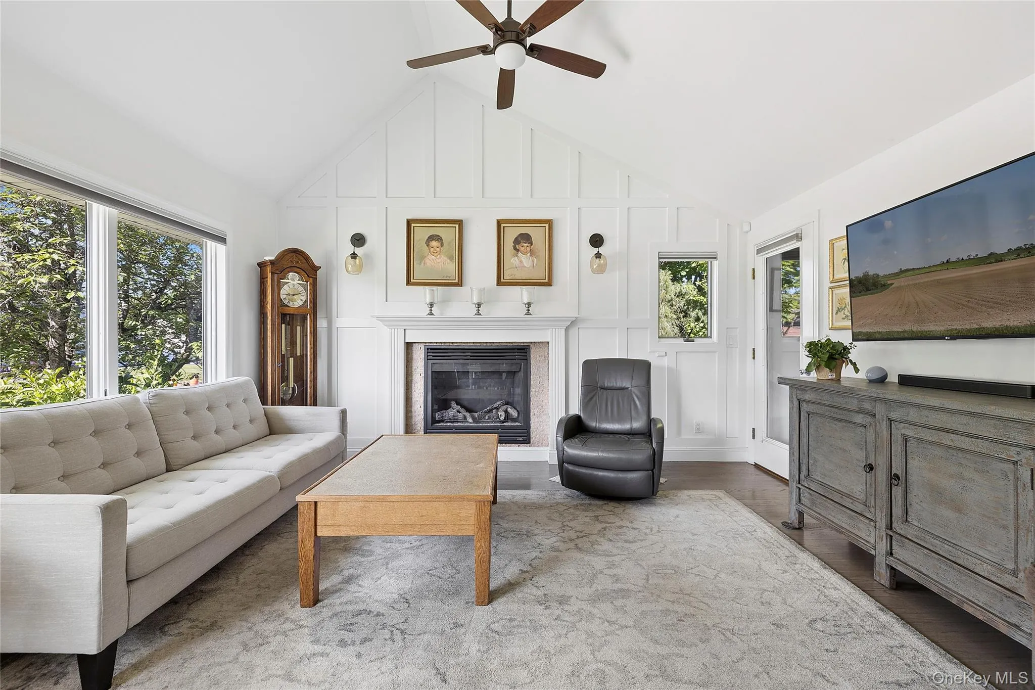 Vaulted ceiling living space featuring a gas fireplace with mantel, board and batten wall detailing, large windows, and dark wood-finish flooring Vaulted ceiling living space featuring a gas fireplace with mantel, board and batten wall detailing, large windows, and dark wood-finish flooring