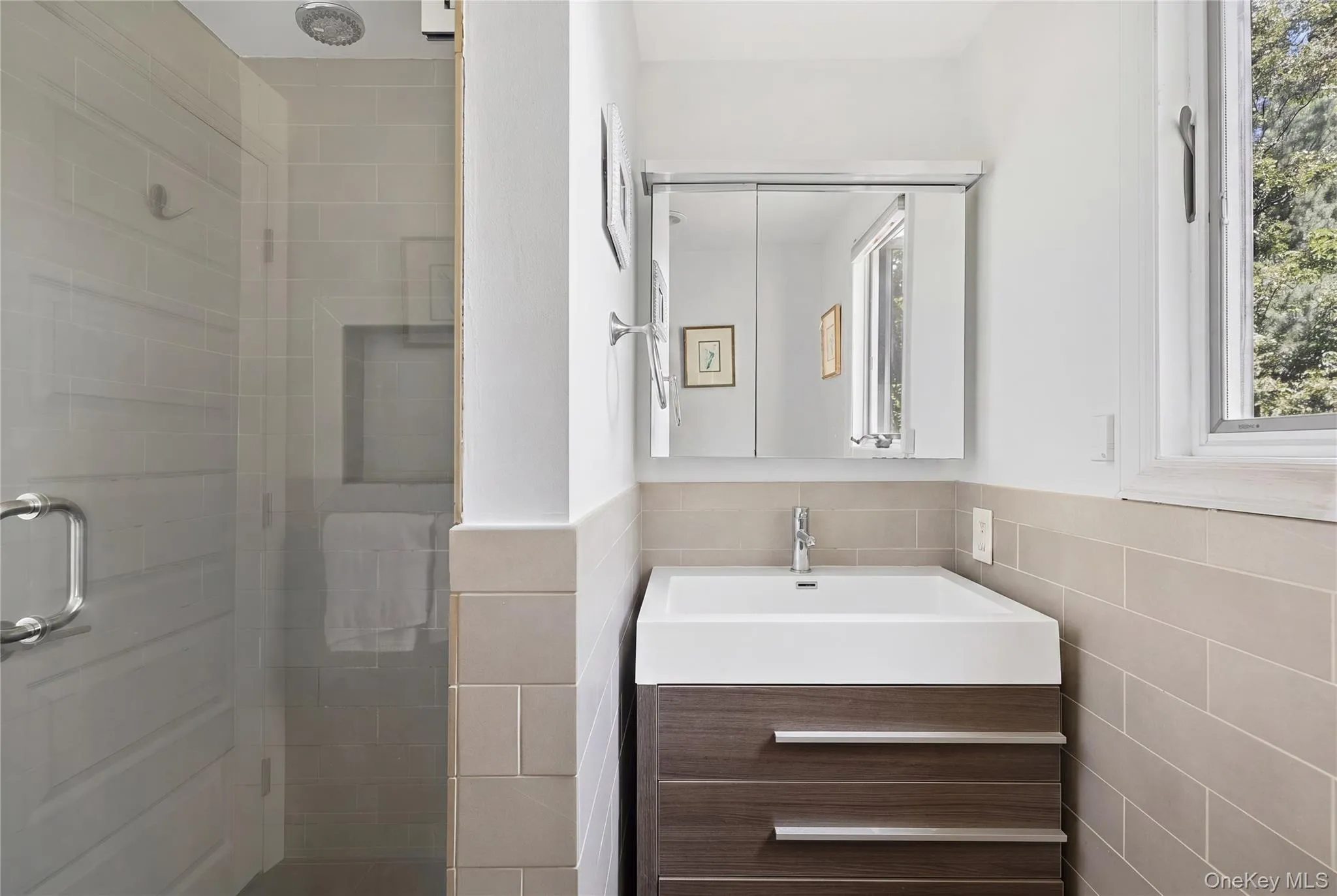 Contemporary bathroom featuring a glass-enclosed shower with a recessed shelf, a rain showerhead, a floating wood-finish vanity with an integrated sink, a mirrored medicine cabinet, and a window providing natural illumination Contemporary bathroom featuring a glass-enclosed shower with a recessed shelf, a rain showerhead, a floating wood-finish vanity with an integrated sink, a mirrored medicine cabinet, and a window providing natural illumination