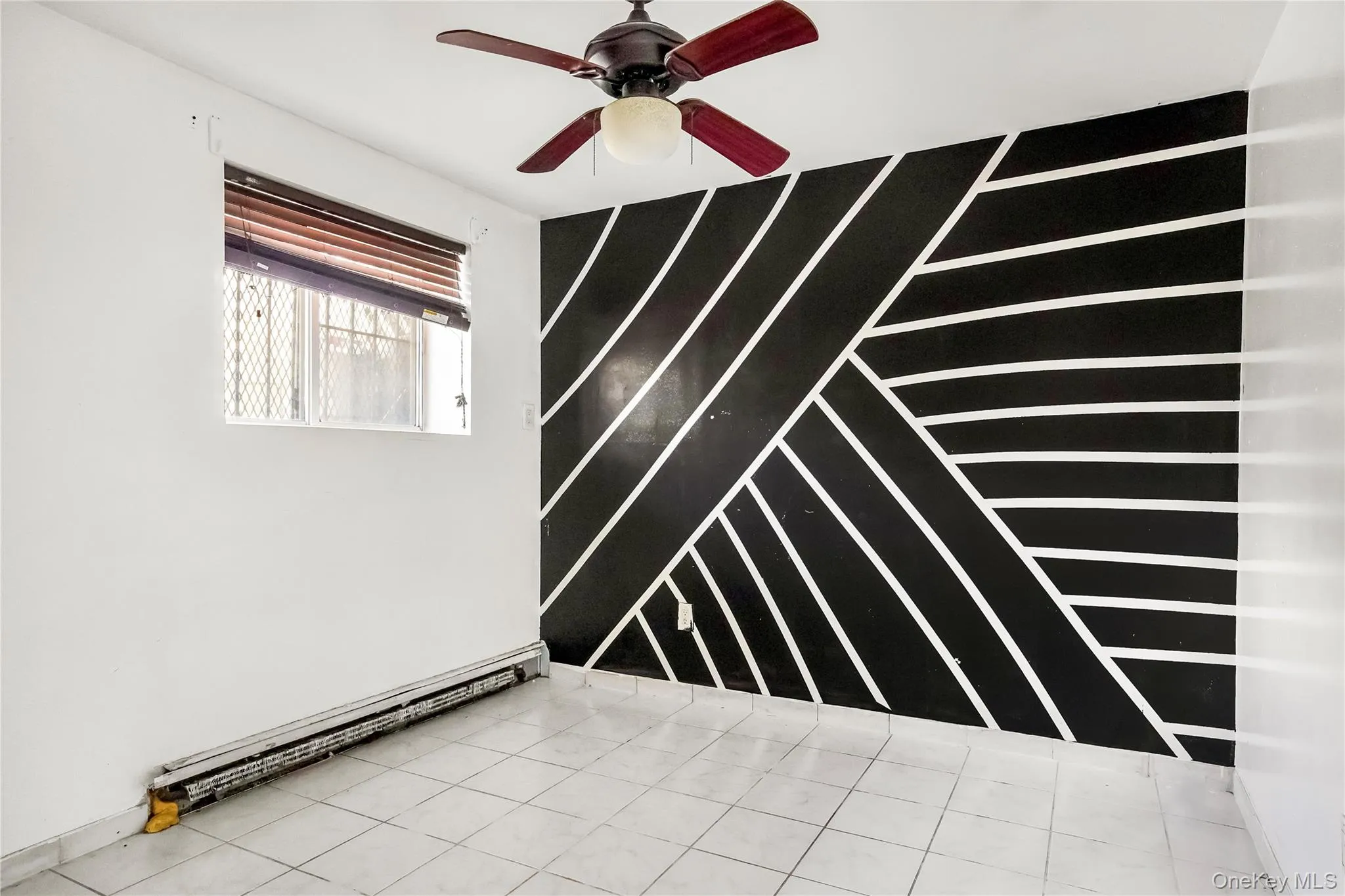 Interior room featuring a black accent wall with white geometric patterns, a ceiling fan with light fixture, a window with horizontal blinds and security bars, baseboard heating, and square tile flooring Interior room featuring a black accent wall with white geometric patterns, a ceiling fan with light fixture, a window with horizontal blinds and security bars, baseboard heating, and square tile flooring