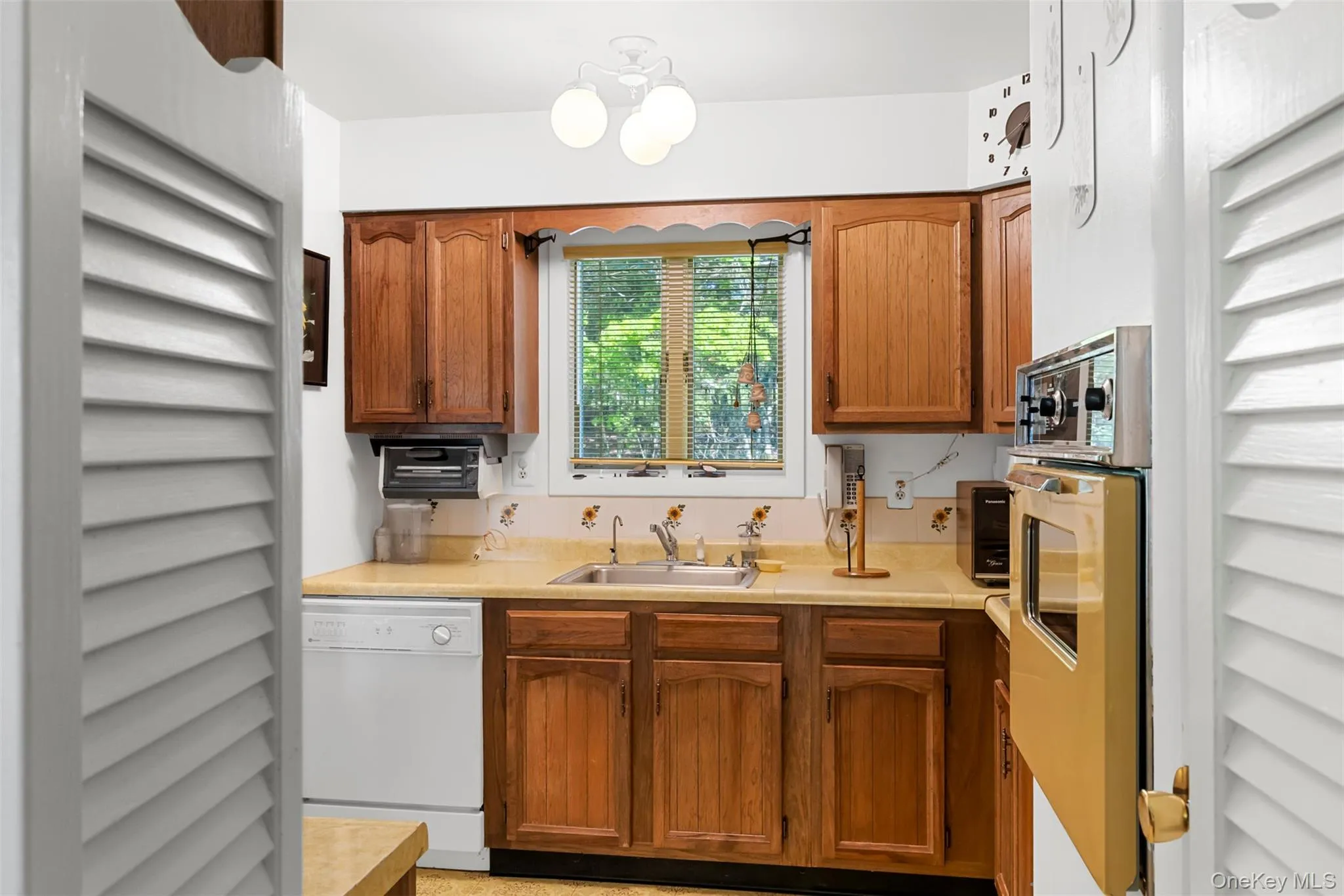 Kitchen with white dishwasher, a sink, oven, light countertops, and brown cabinetry Kitchen with white dishwasher, a sink, oven, light countertops, and brown cabinetry