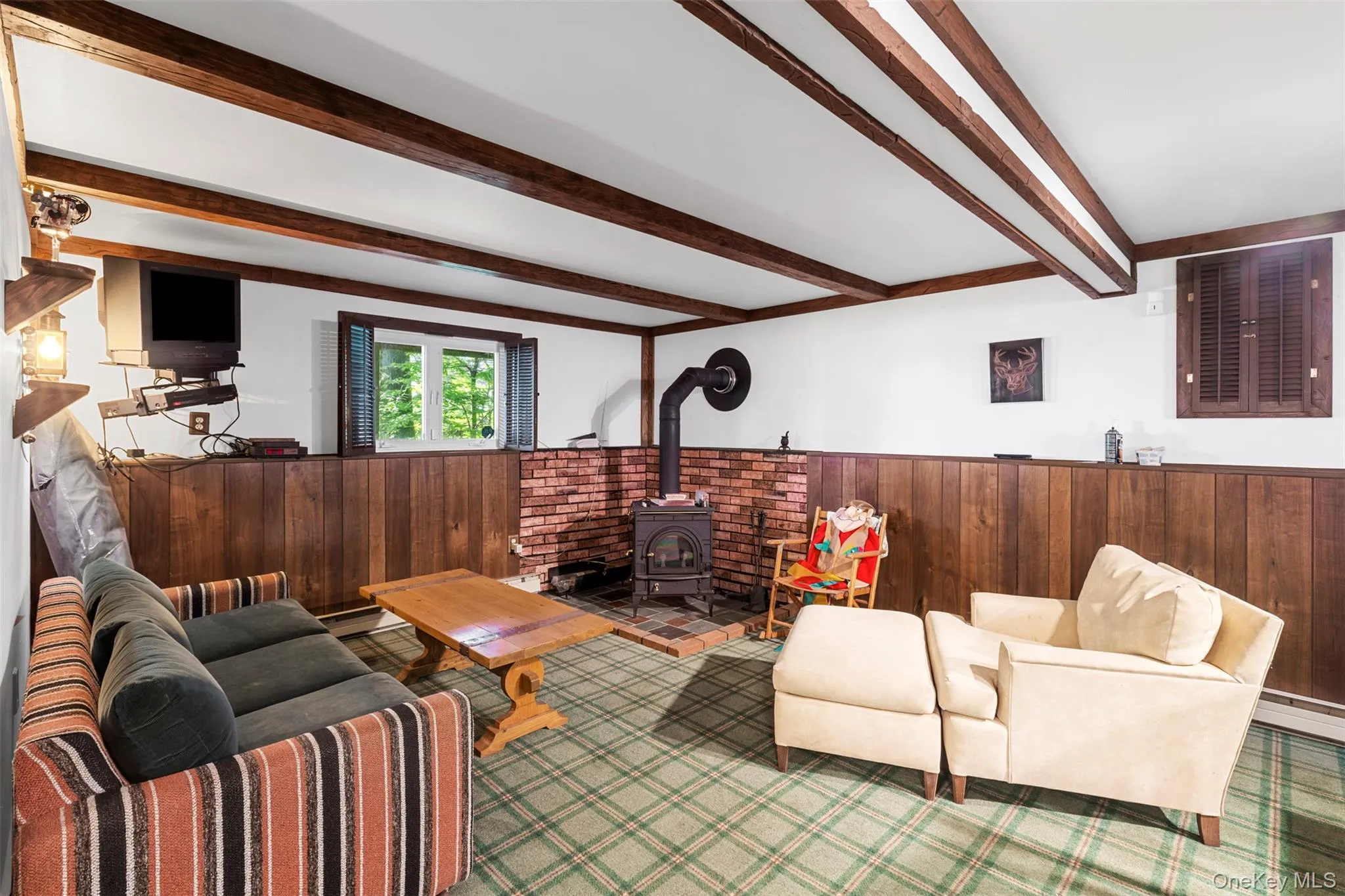 Living area featuring wooden walls, wainscoting, beamed ceiling, a wood stove, and a baseboard radiator Living area featuring wooden walls, wainscoting, beamed ceiling, a wood stove, and a baseboard radiator