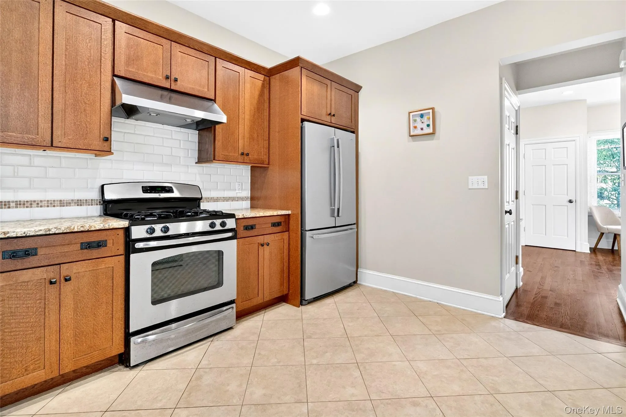 Kitchen featuring extensive wood cabinetry, stainless steel appliances, a white subway tile backsplash with decorative accent, stone countertops, and neutral-toned floor tiling Kitchen featuring extensive wood cabinetry, stainless steel appliances, a white subway tile backsplash with decorative accent, stone countertops, and neutral-toned floor tiling