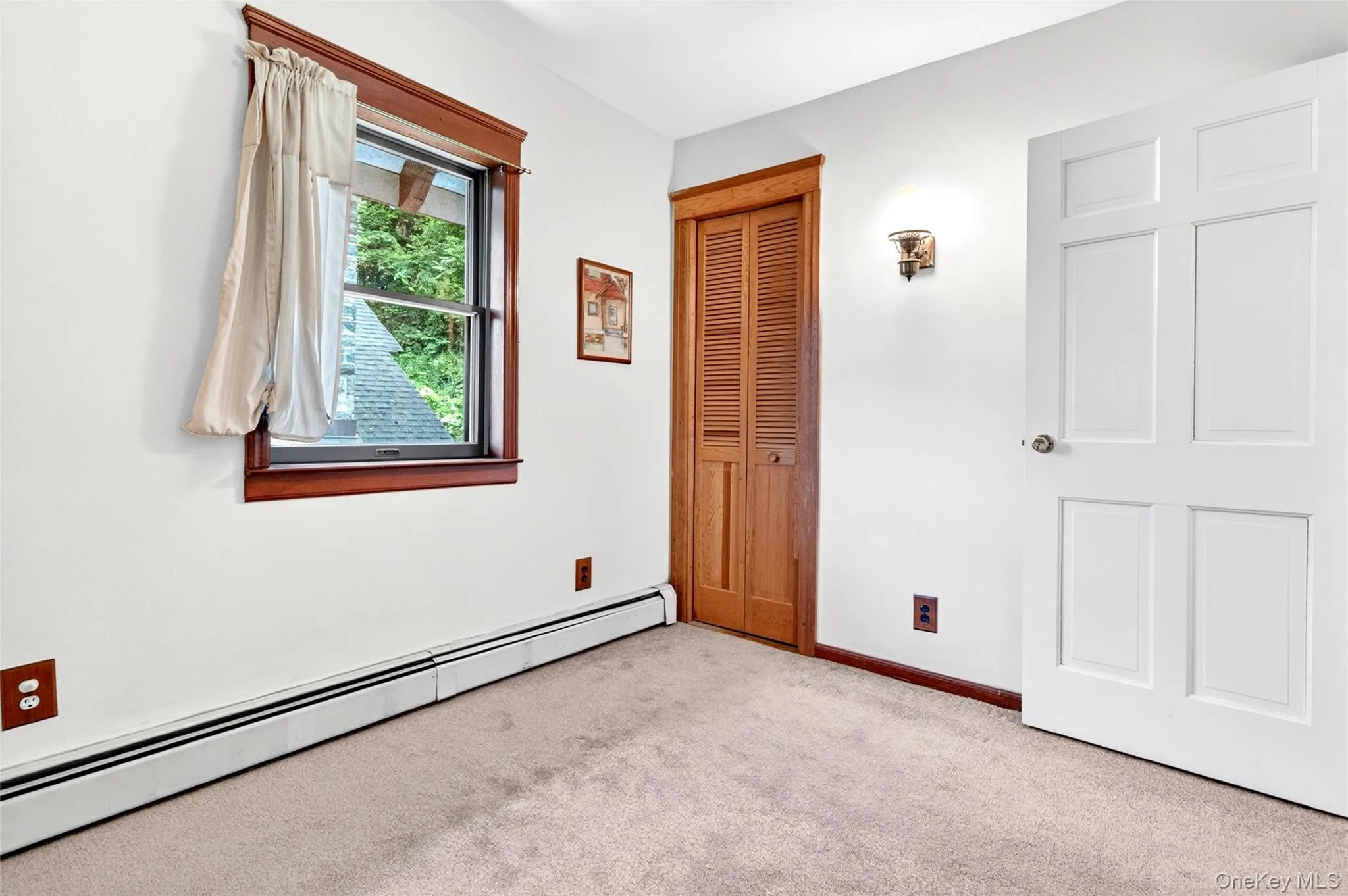 Carpeted room featuring a double-hung window with wood trim, a paneled door, louvered bi-fold closet doors, and baseboard heating Carpeted room featuring a double-hung window with wood trim, a paneled door, louvered bi-fold closet doors, and baseboard heating