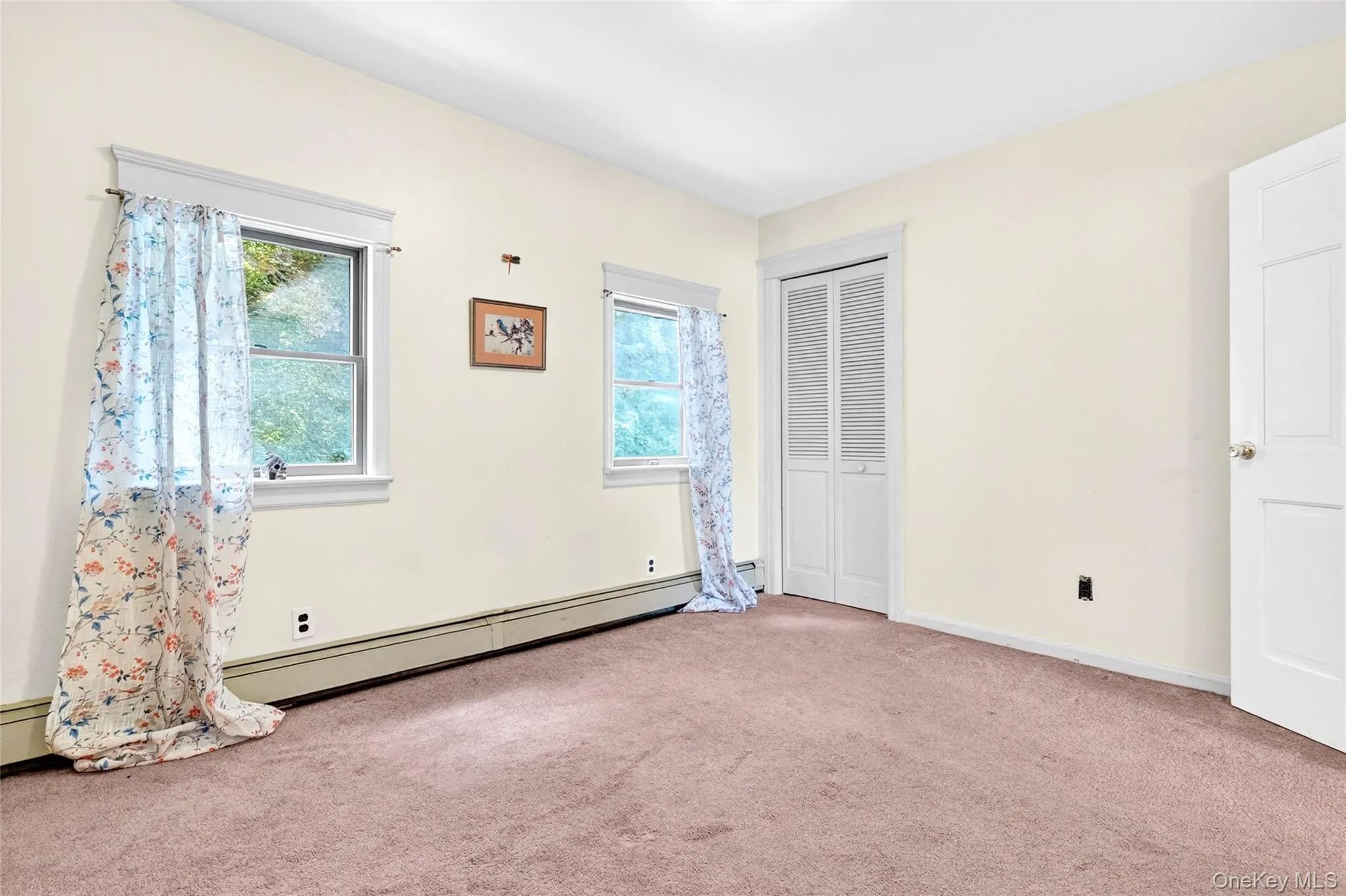Carpeted interior with two double-hung windows, a bi-fold louvered closet door, and baseboard heating Carpeted interior with two double-hung windows, a bi-fold louvered closet door, and baseboard heating
