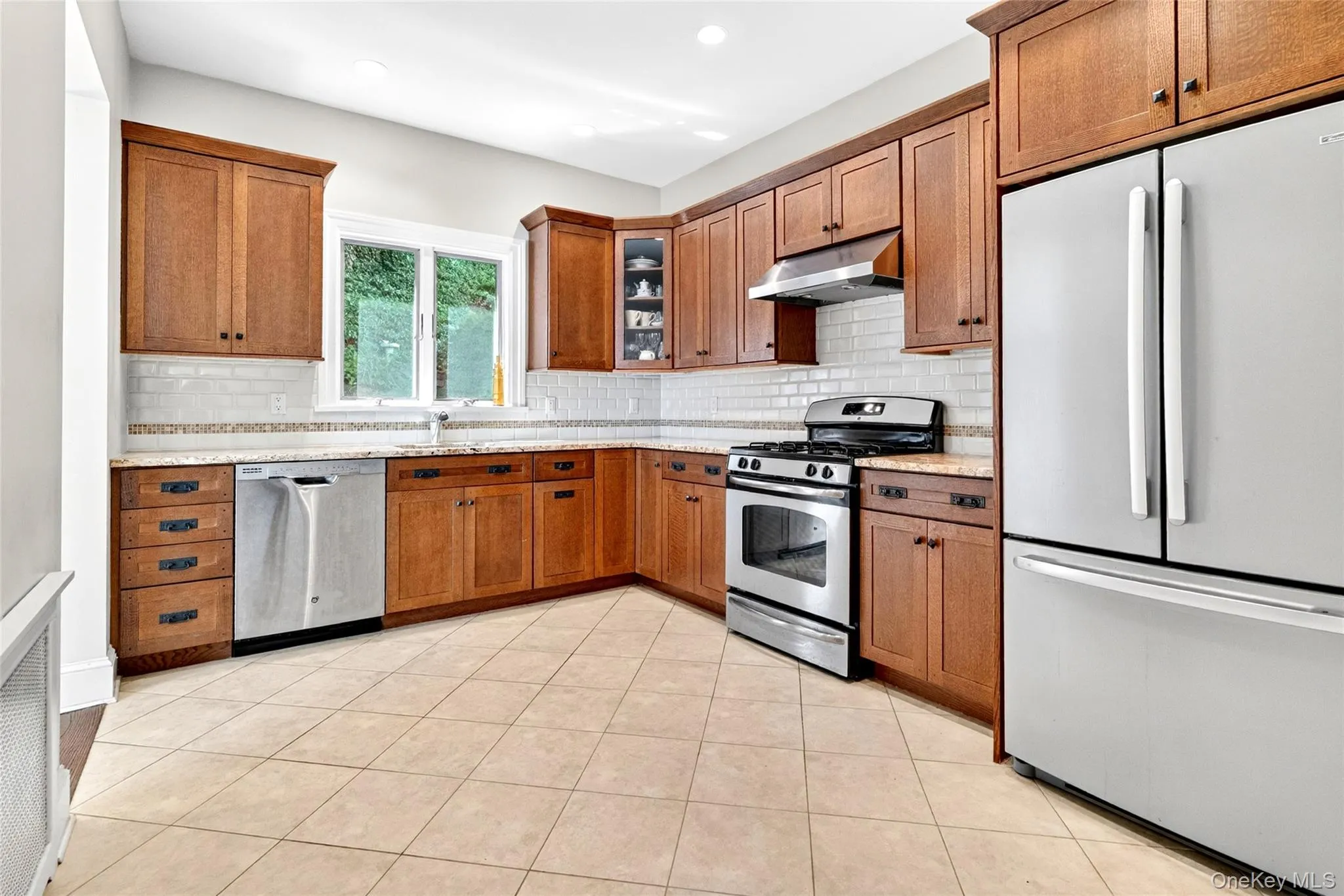 Kitchen featuring wood cabinetry, stainless steel appliances, a tile backsplash, and light-toned tile flooring Kitchen featuring wood cabinetry, stainless steel appliances, a tile backsplash, and light-toned tile flooring