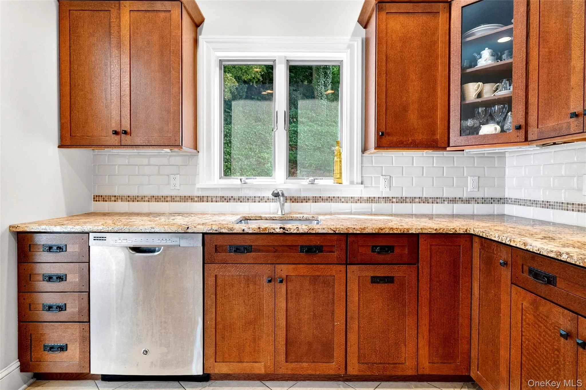 Kitchen featuring wood cabinetry, solid surface countertops, white subway tile backsplash with a decorative accent band, stainless steel dishwasher, and a window above the sink Kitchen featuring wood cabinetry, solid surface countertops, white subway tile backsplash with a decorative accent band, stainless steel dishwasher, and a window above the sink