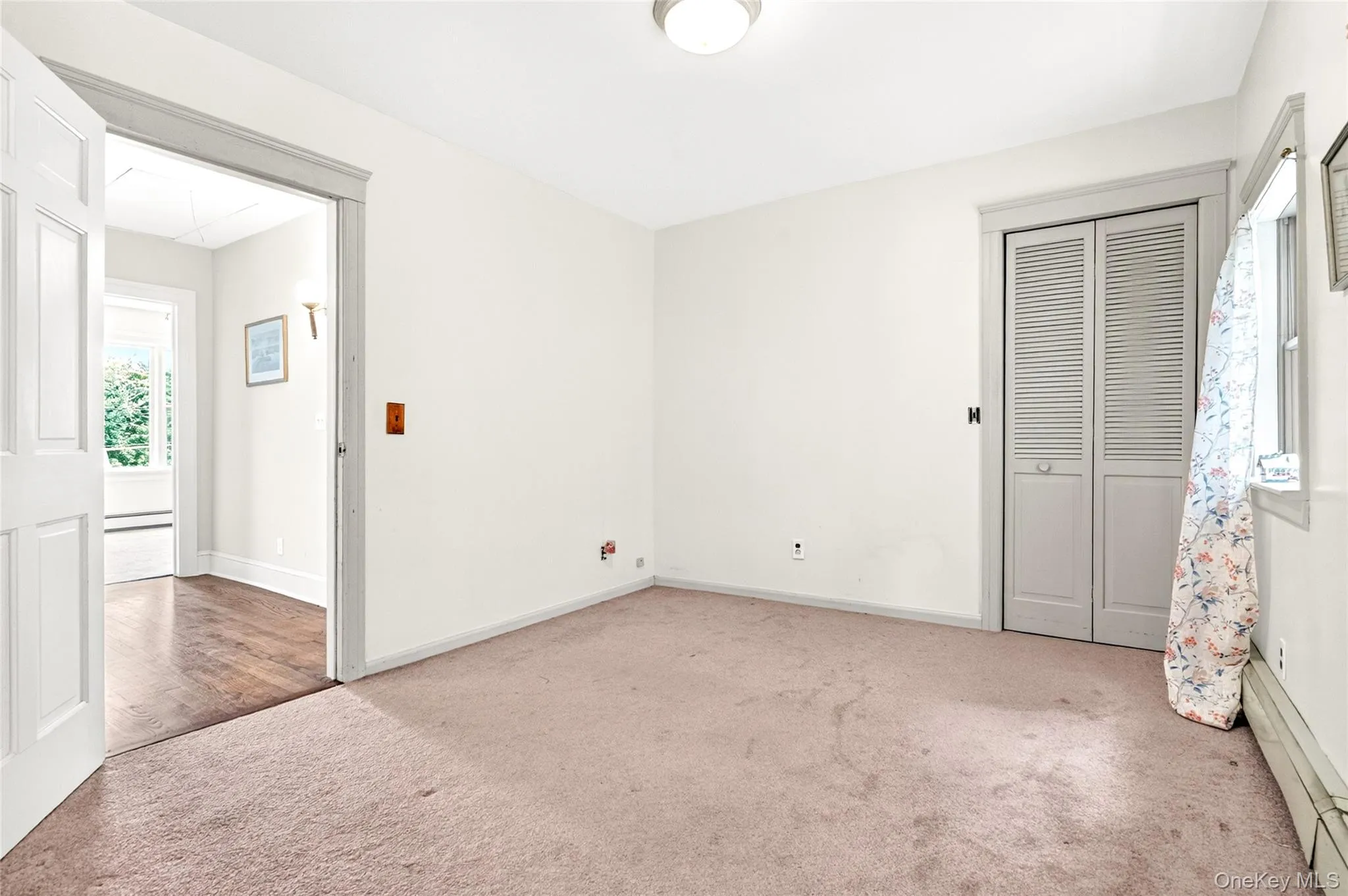 Carpeted room with a bi-fold louvered closet door, a window, and a flush-mount ceiling light Carpeted room with a bi-fold louvered closet door, a window, and a flush-mount ceiling light