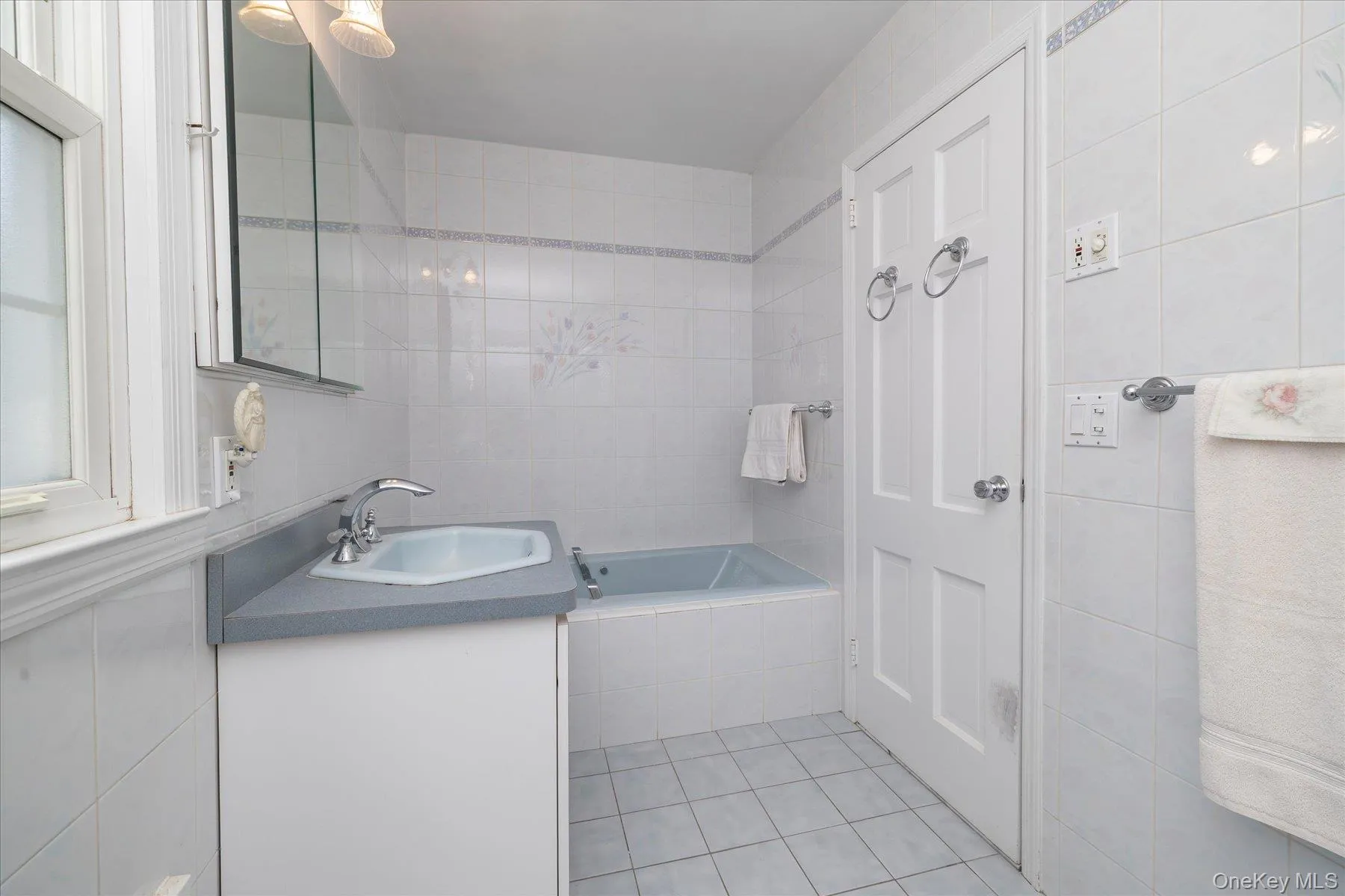 Bathroom featuring a built-in bathtub, a window with trim, a vanity with an integrated sink, a mirrored medicine cabinet, and tiled walls and flooring. Bathroom featuring a built-in bathtub, a window with trim, a vanity with an integrated sink, a mirrored medicine cabinet, and tiled walls and flooring.