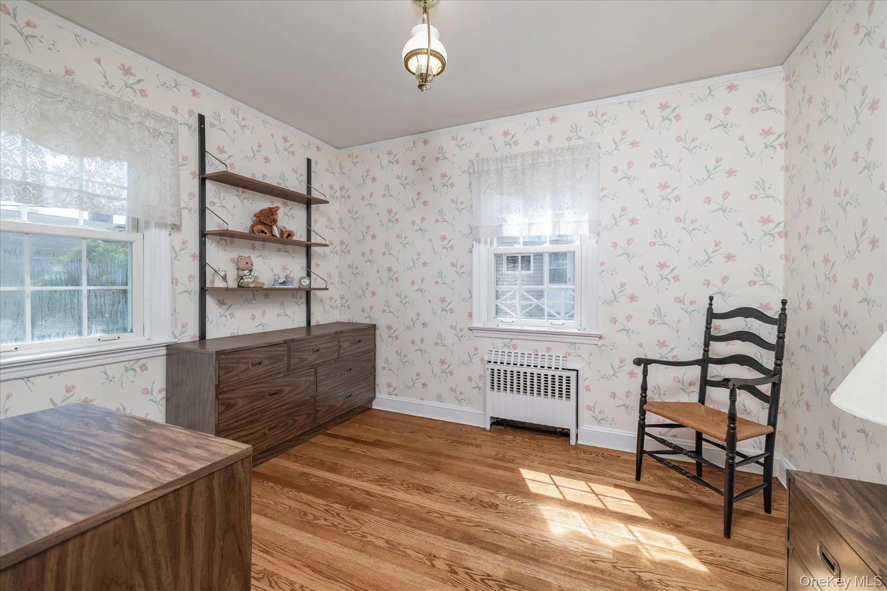Room featuring wood-finish flooring, floral pattern wallpaper, and two windows with white trim. Room featuring wood-finish flooring, floral pattern wallpaper, and two windows with white trim.