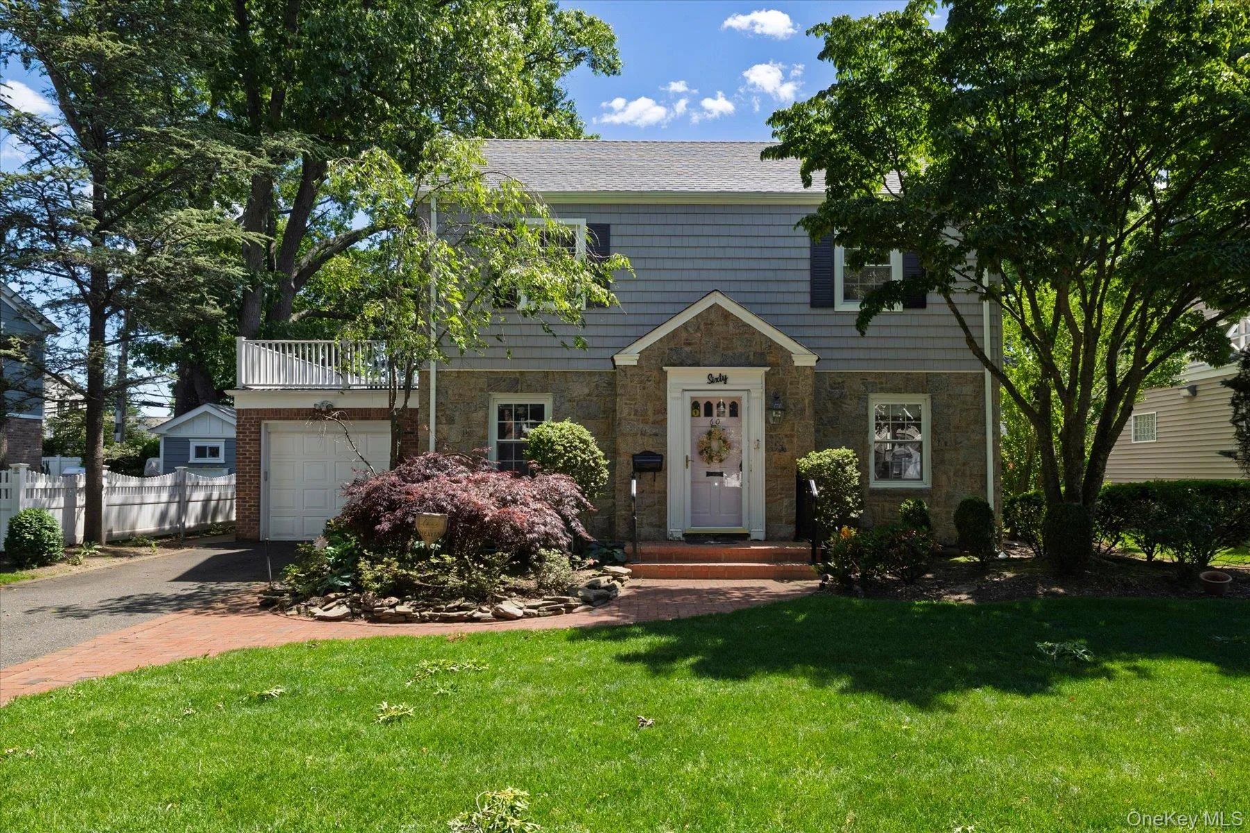 Traditional two-story residence featuring a stone and shingle exterior, an integrated garage with an overhead balcony, a paved driveway, and mature trees framing the property Traditional two-story residence featuring a stone and shingle exterior, an integrated garage with an overhead balcony, a paved driveway, and mature trees framing the property