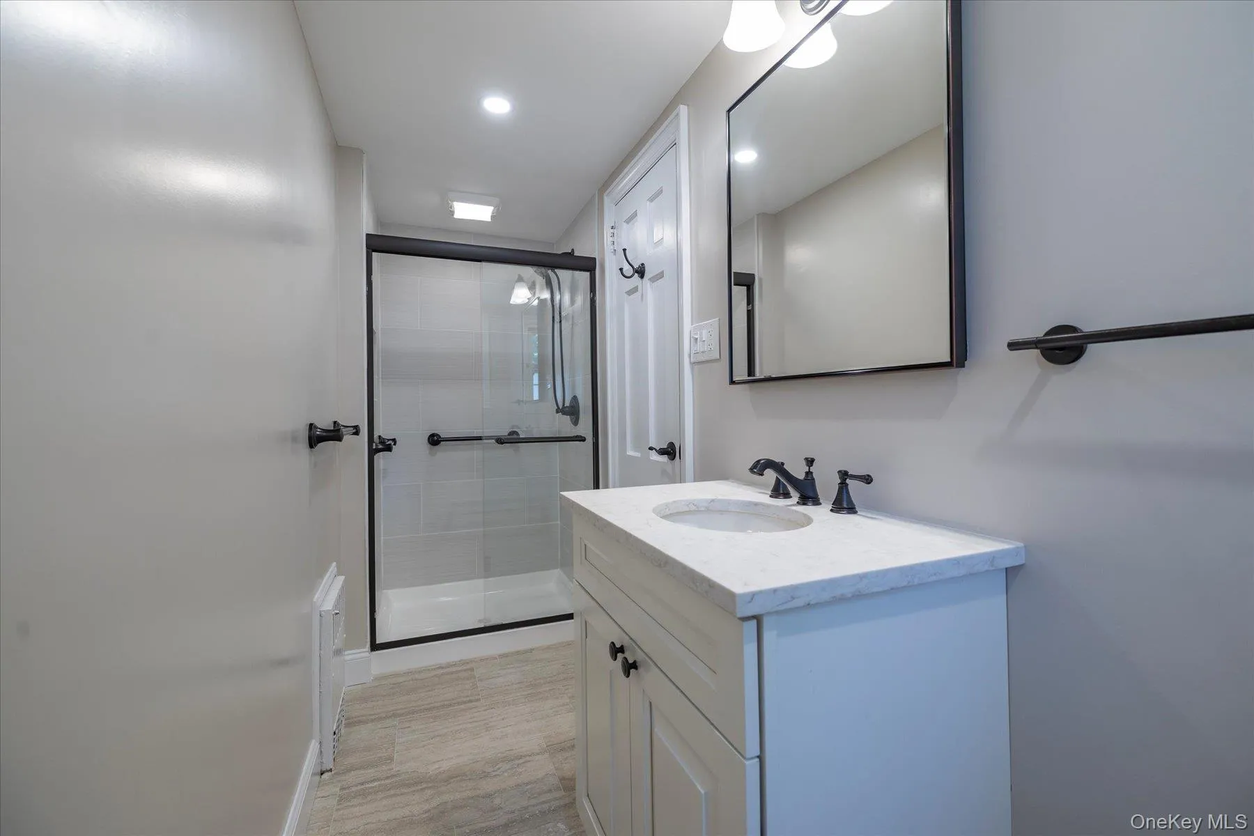 Bathroom featuring a glass-enclosed shower with black trim, a vanity with a white countertop and integrated sink, a rectangular framed mirror, and wood-finish flooring. Bathroom featuring a glass-enclosed shower with black trim, a vanity with a white countertop and integrated sink, a rectangular framed mirror, and wood-finish flooring.