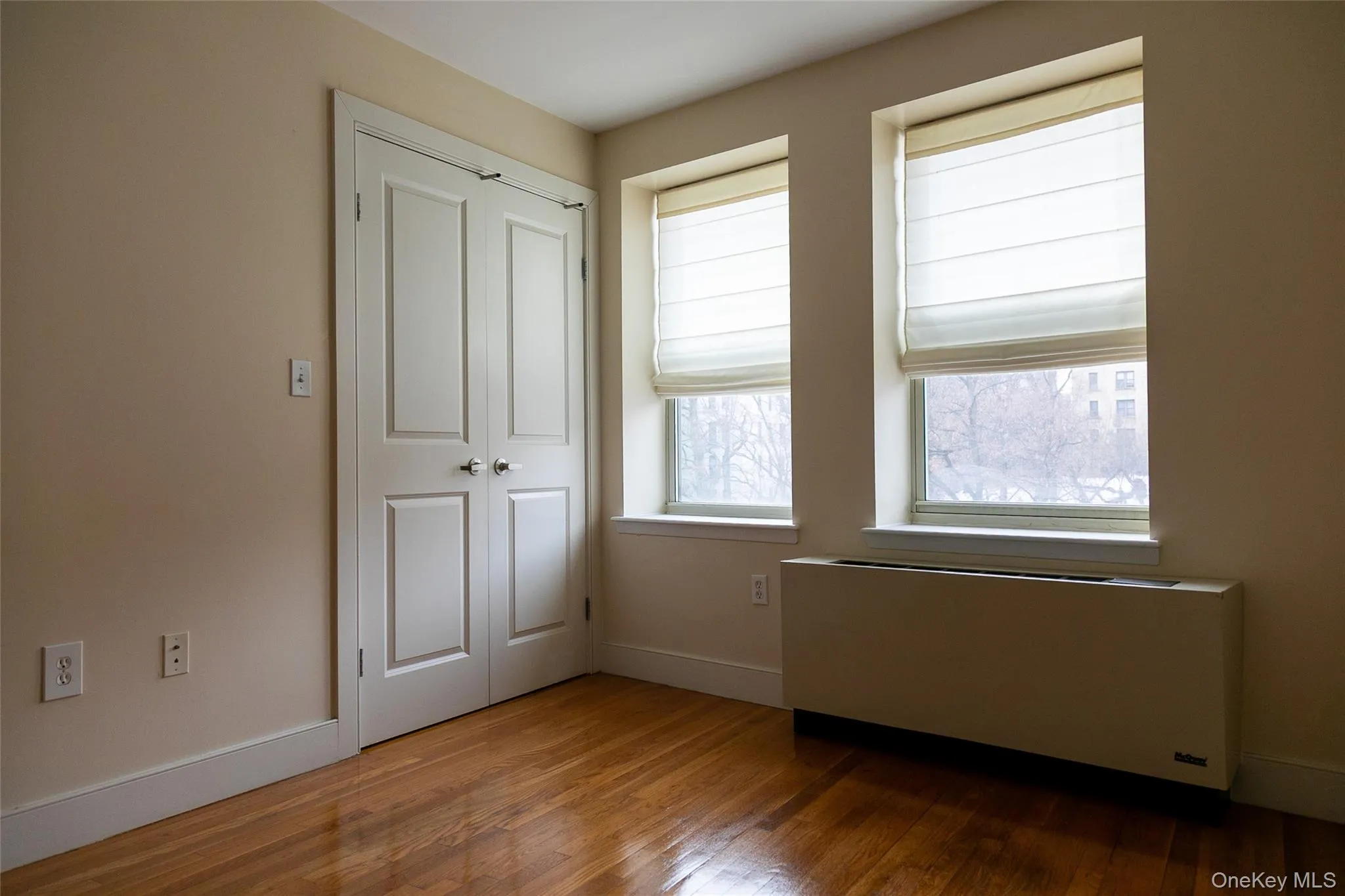 Hardwood flooring throughout, featuring two windows with privacy shades, a built-in heating unit, and a double-door closet Hardwood flooring throughout, featuring two windows with privacy shades, a built-in heating unit, and a double-door closet