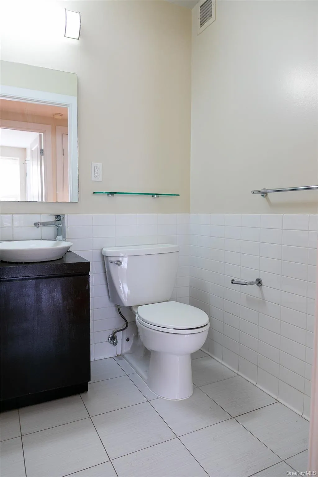 Bathroom featuring a wall-mounted mirror, vessel sink, dark wood vanity, white toilet, and white tile flooring Bathroom featuring a wall-mounted mirror, vessel sink, dark wood vanity, white toilet, and white tile flooring