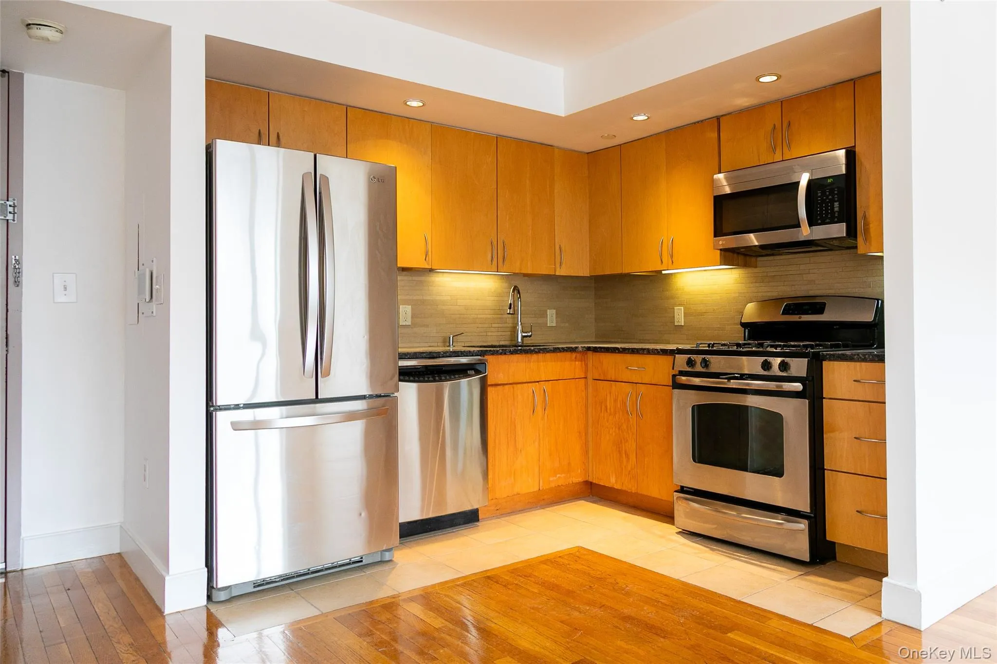 Modern kitchen featuring stainless steel appliances, light wood-finish cabinetry, granite-style countertops, and a tiled backsplash Modern kitchen featuring stainless steel appliances, light wood-finish cabinetry, granite-style countertops, and a tiled backsplash