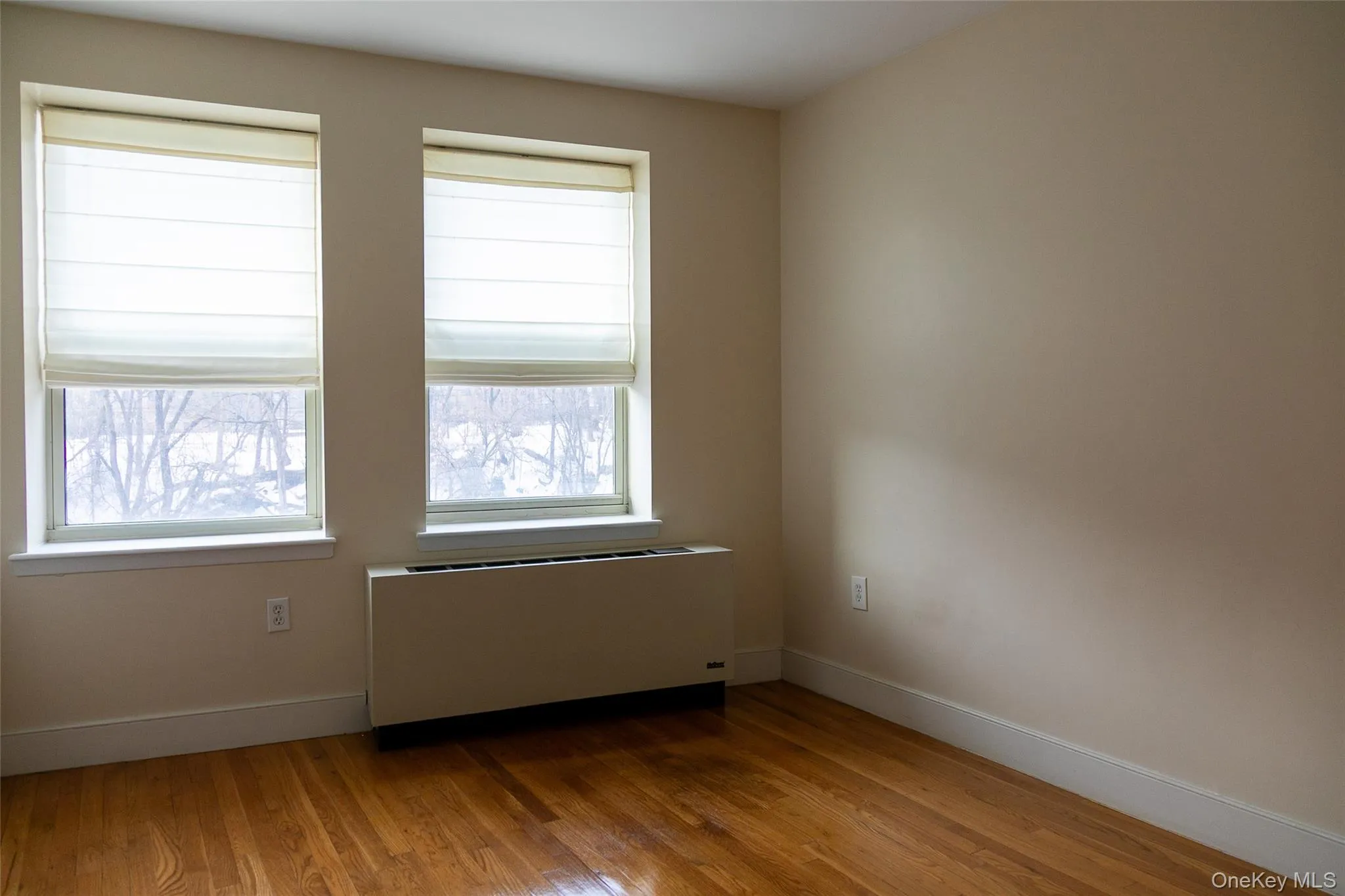 Room featuring two windows with privacy shades, hardwood flooring, and neutral painted walls Room featuring two windows with privacy shades, hardwood flooring, and neutral painted walls