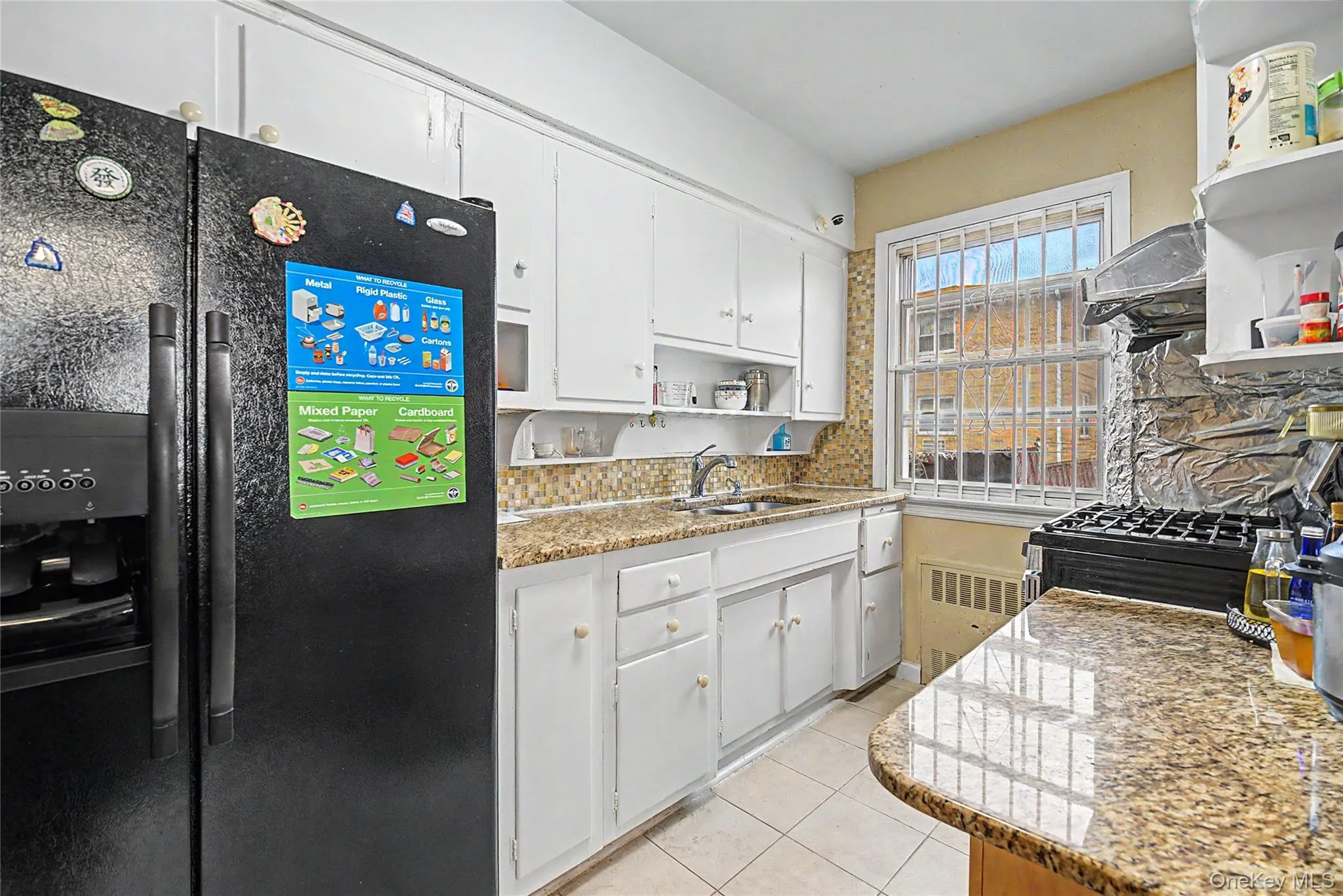 Compact kitchen featuring white cabinetry, a black side-by-side refrigerator, speckled granite-finish countertops, mosaic tile backsplash, and a double-hung window Compact kitchen featuring white cabinetry, a black side-by-side refrigerator, speckled granite-finish countertops, mosaic tile backsplash, and a double-hung window