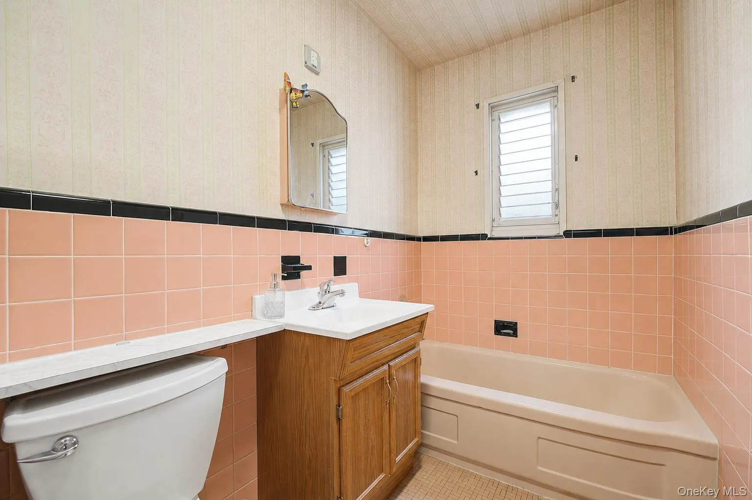 Bathroom featuring peach-toned wall tile with black trim, a wood-finish vanity with a white countertop and integrated sink, a white toilet, and a window with horizontal slat blinds Bathroom featuring peach-toned wall tile with black trim, a wood-finish vanity with a white countertop and integrated sink, a white toilet, and a window with horizontal slat blinds