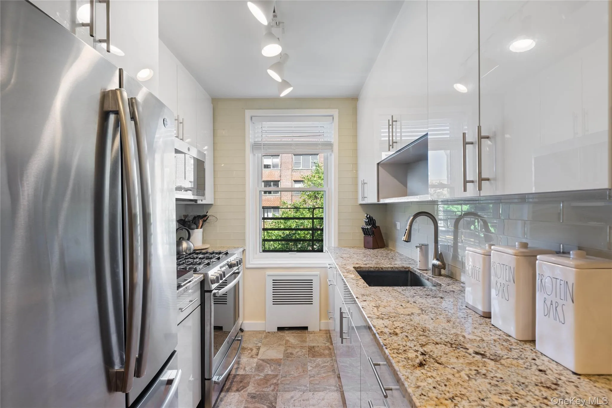 Galley kitchen featuring stainless steel appliances, light-toned granite countertops, white high-gloss cabinetry, and a subway tile backsplash Galley kitchen featuring stainless steel appliances, light-toned granite countertops, white high-gloss cabinetry, and a subway tile backsplash