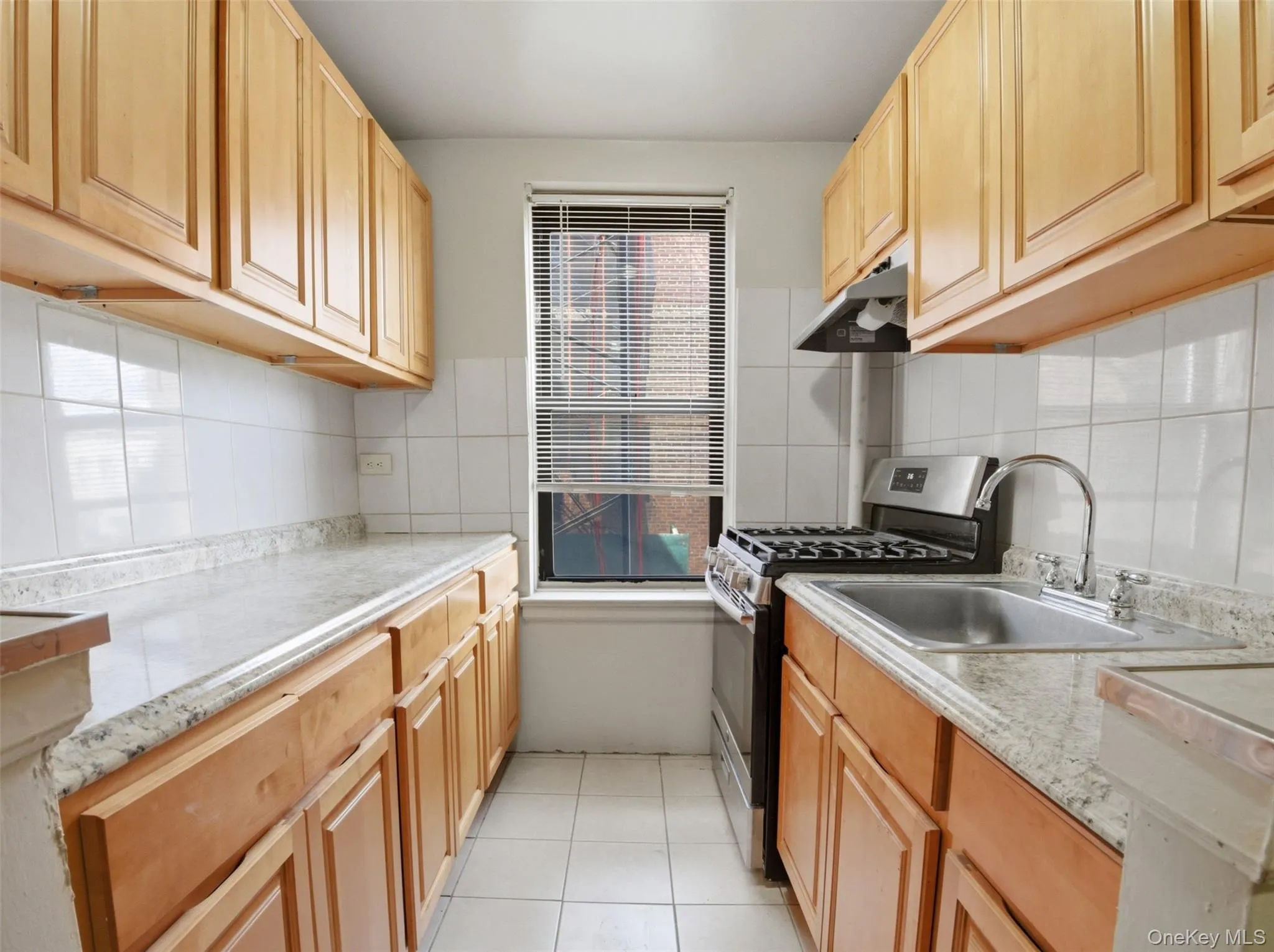 Galley kitchen featuring wood-finish cabinetry, light-toned countertops, white tiled backsplash, stainless steel sink, and gas range with coordinating range hood Galley kitchen featuring wood-finish cabinetry, light-toned countertops, white tiled backsplash, stainless steel sink, and gas range with coordinating range hood