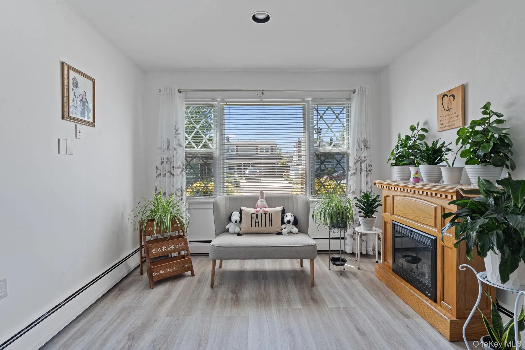 Bright living space featuring a large window with white blinds, light wood-finish flooring, and a built-in fireplace with a light wood mantle Bright living space featuring a large window with white blinds, light wood-finish flooring, and a built-in fireplace with a light wood mantle