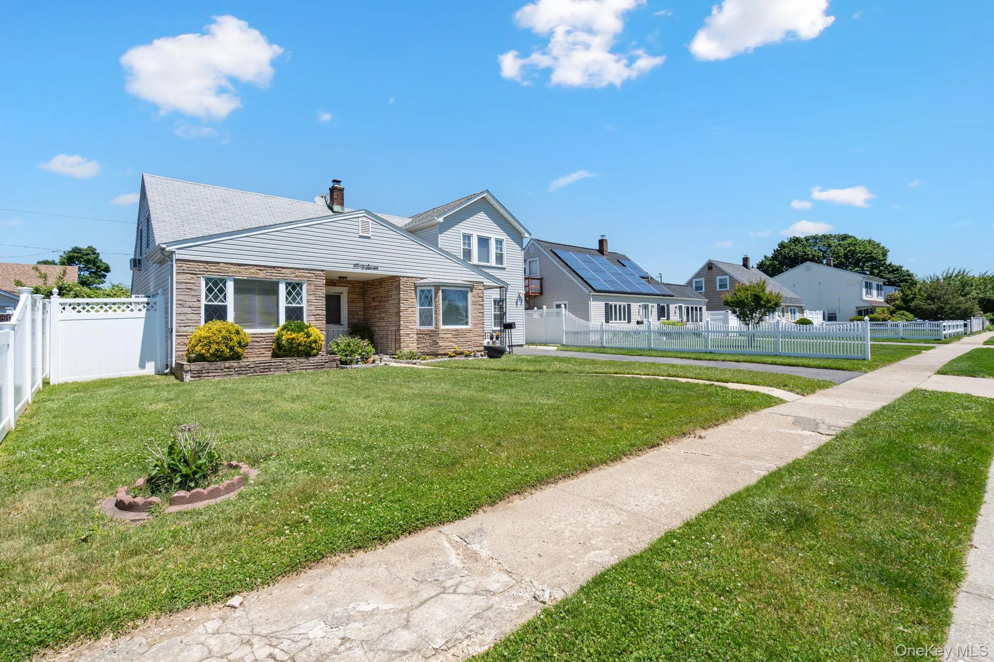 Stone and siding exterior featuring a pitched roofline, front landscaping, and a white privacy fence Stone and siding exterior featuring a pitched roofline, front landscaping, and a white privacy fence