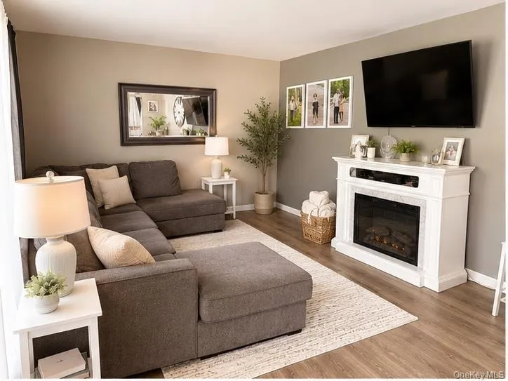Living area featuring vinyl-finish flooring, a white mantel with an electric fireplace insert, and a large wall-mounted television Living area featuring vinyl-finish flooring, a white mantel with an electric fireplace insert, and a large wall-mounted television