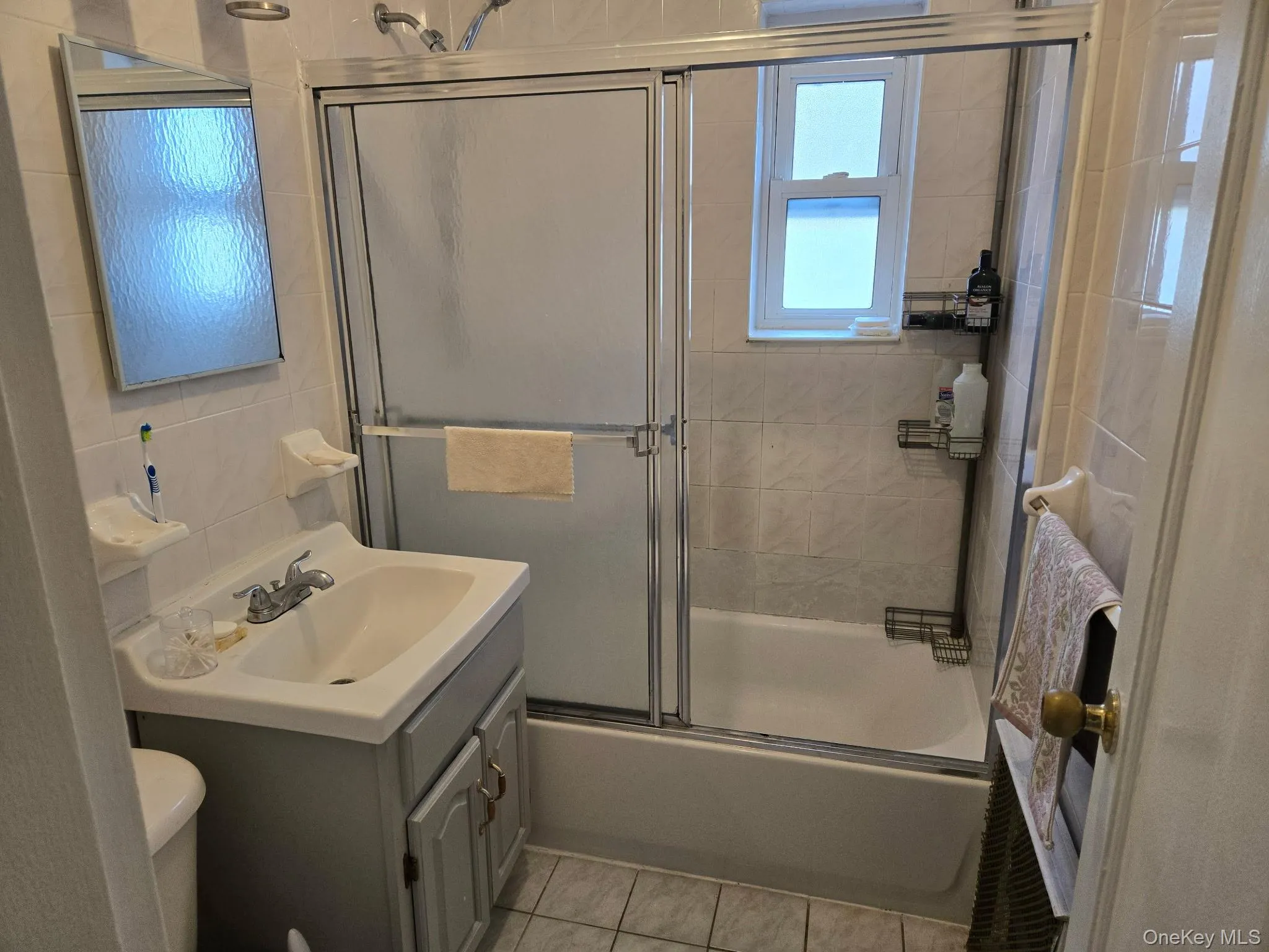 Full bathroom featuring a white porcelain tub with sliding glass shower doors, a window above the tub, a vanity with a drop-in sink, and a wall-mounted mirror Full bathroom featuring a white porcelain tub with sliding glass shower doors, a window above the tub, a vanity with a drop-in sink, and a wall-mounted mirror