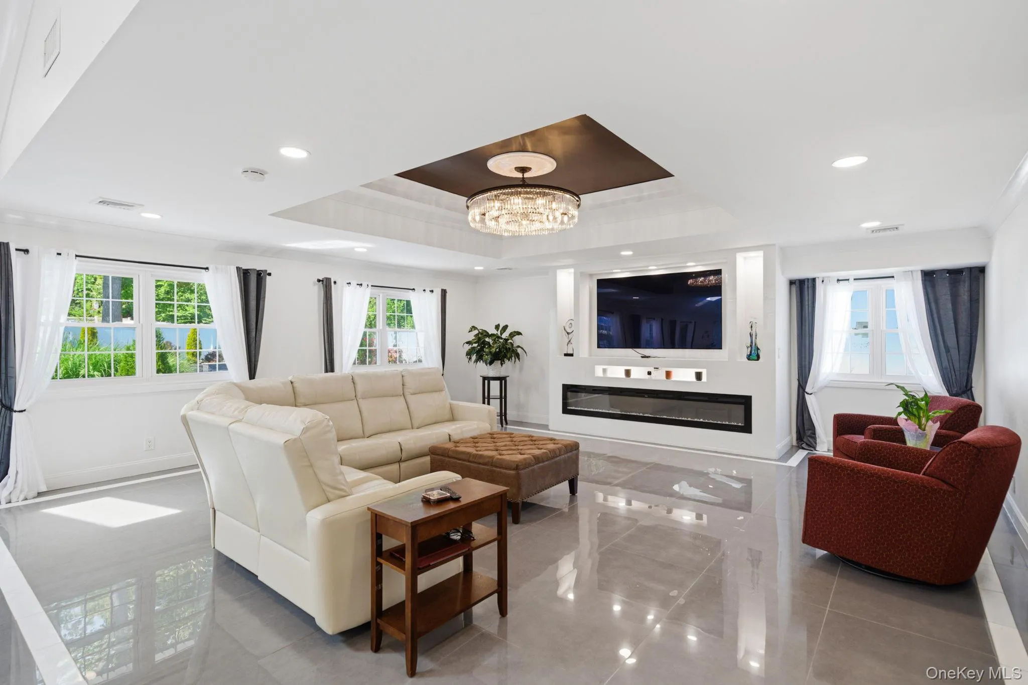 Open and bright living room with decorative ceiling, stunning chandelier, recessed lighting, wall of windows, electric fireplace and porcelain tile floors. Open and bright living room with decorative ceiling, stunning chandelier, recessed lighting, wall of windows, electric fireplace and porcelain tile floors.