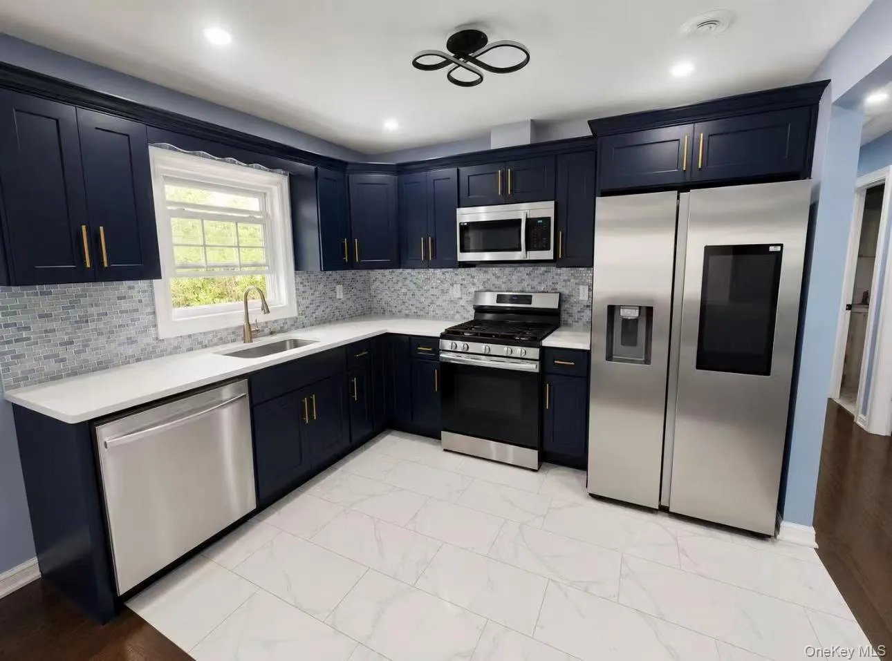 Modern kitchen featuring navy cabinetry with brass hardware, white countertops, a mosaic tile backsplash, stainless steel appliances, and white tile flooring Modern kitchen featuring navy cabinetry with brass hardware, white countertops, a mosaic tile backsplash, stainless steel appliances, and white tile flooring