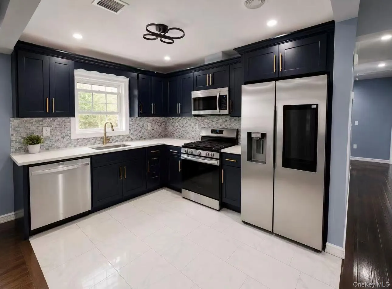 Modern kitchen featuring dark blue cabinetry with gold-finish hardware, stainless steel appliances, white countertops, a tiled backsplash, and white tile flooring Modern kitchen featuring dark blue cabinetry with gold-finish hardware, stainless steel appliances, white countertops, a tiled backsplash, and white tile flooring