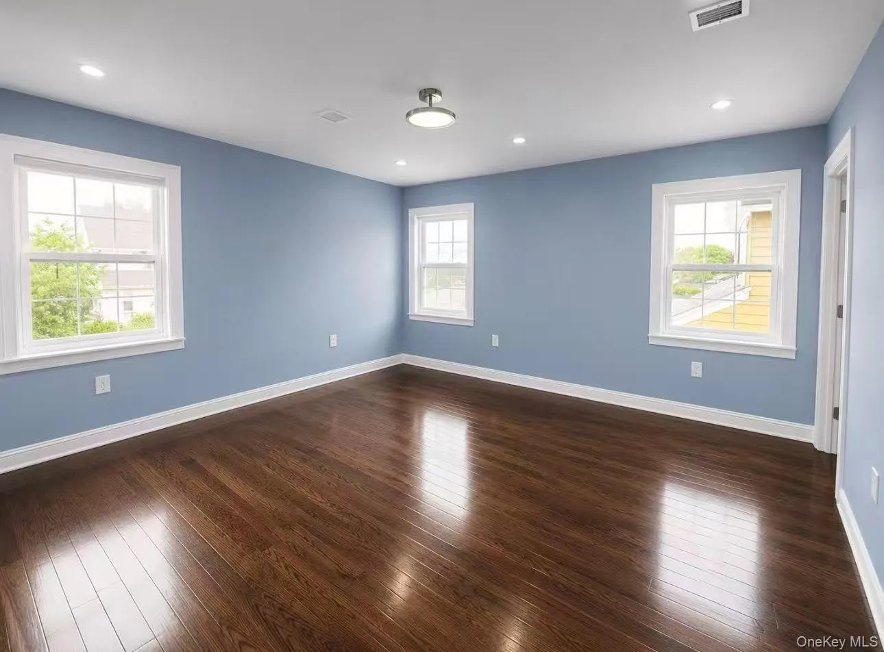 Spacious room featuring rich wood-finish flooring, multiple windows with white trim, recessed lighting, a central flush-mount light fixture, and crisp white baseboards Spacious room featuring rich wood-finish flooring, multiple windows with white trim, recessed lighting, a central flush-mount light fixture, and crisp white baseboards