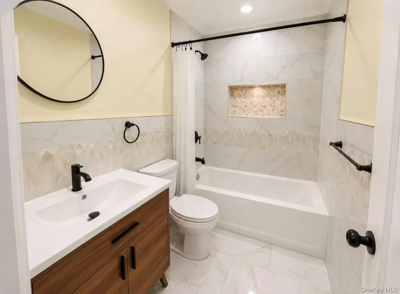 Bathroom featuring a wood-finish vanity with an integrated white sink, matte black fixtures, a white bathtub with a tiled surround, a recessed shower niche with accent tile, and a circular wall mirror Bathroom featuring a wood-finish vanity with an integrated white sink, matte black fixtures, a white bathtub with a tiled surround, a recessed shower niche with accent tile, and a circular wall mirror