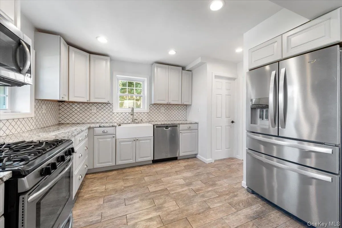 Bright kitchen featuring light cabinetry, stainless steel appliances, a white farmhouse sink, patterned tile backsplash, and wood-finish flooring Bright kitchen featuring light cabinetry, stainless steel appliances, a white farmhouse sink, patterned tile backsplash, and wood-finish flooring