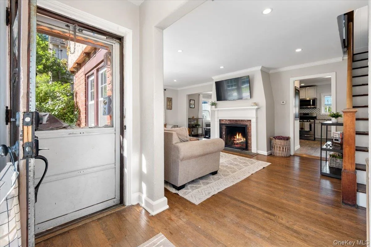 Foyer entry opening to a living area with warm wood-finish flooring, a white mantel fireplace with brick surround, recessed lighting, and a doorway leading to a kitchen Foyer entry opening to a living area with warm wood-finish flooring, a white mantel fireplace with brick surround, recessed lighting, and a doorway leading to a kitchen
