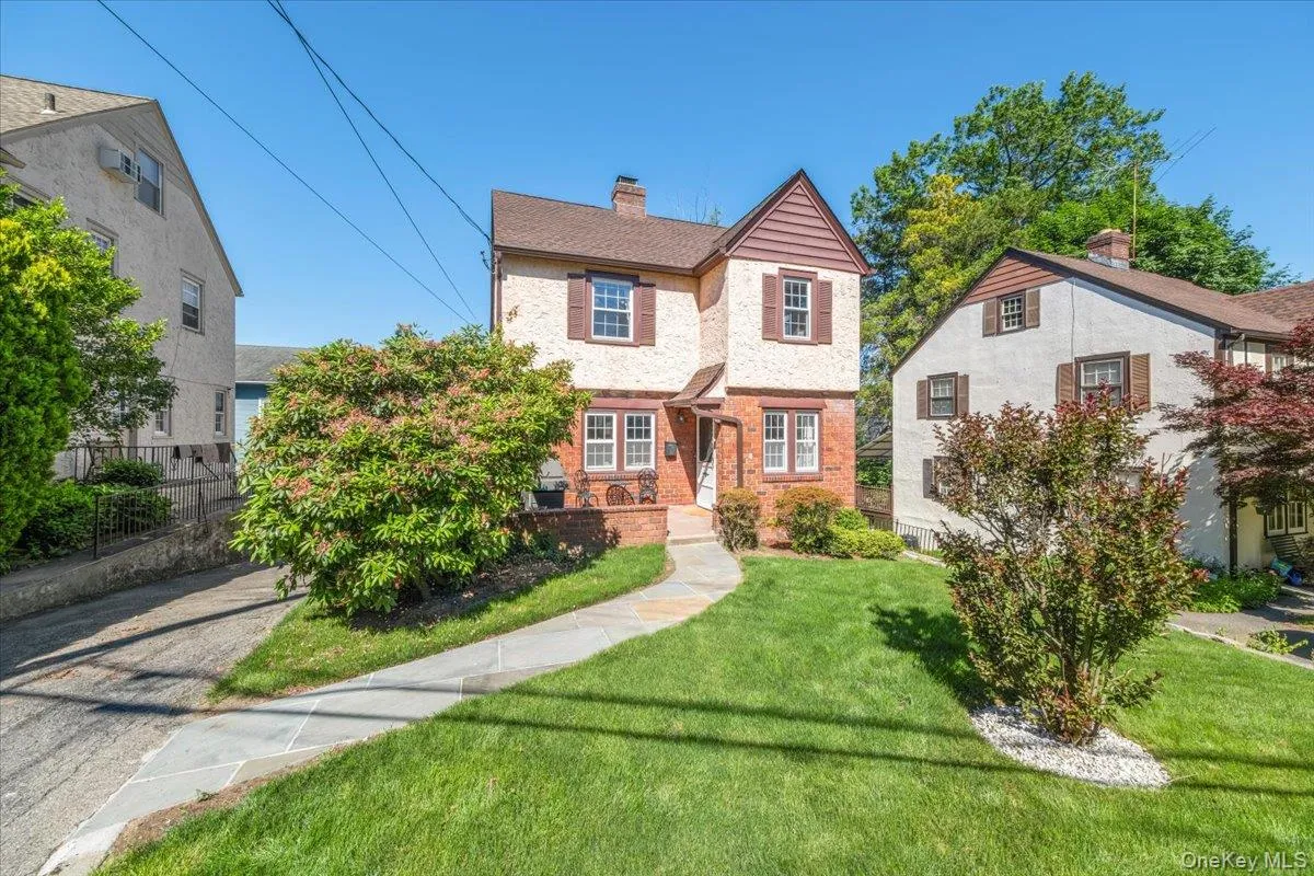 Charming stucco and brick residence featuring a brown shingle roof, dark wood shutters, a stone pathway, and mature landscaping Charming stucco and brick residence featuring a brown shingle roof, dark wood shutters, a stone pathway, and mature landscaping