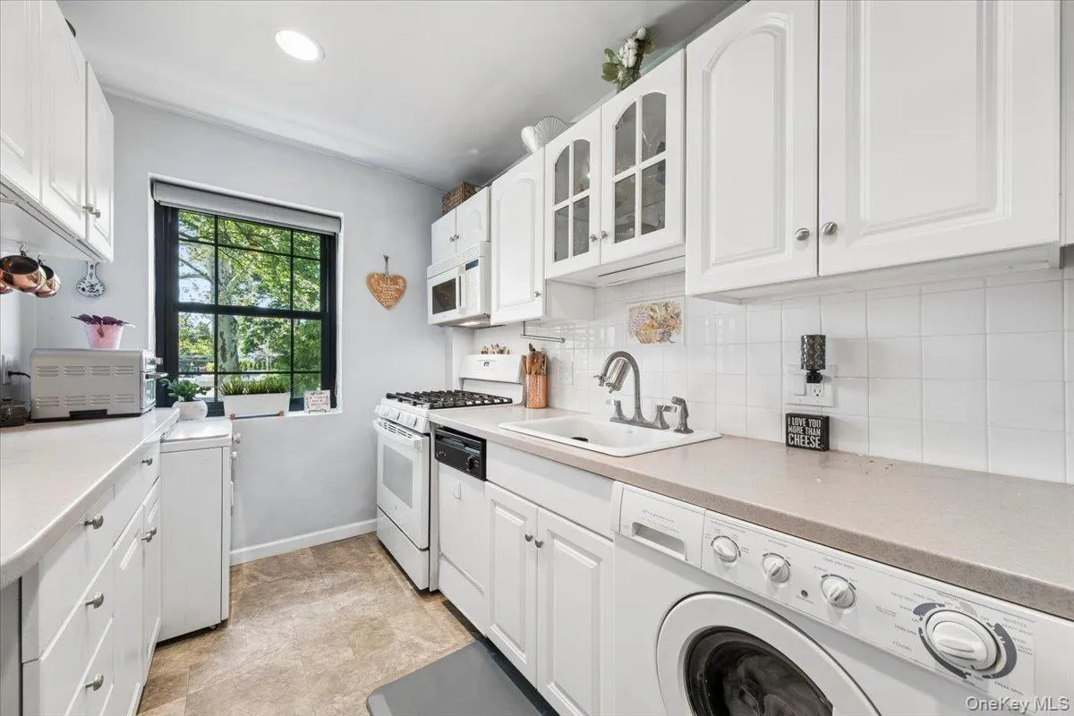 Galley kitchen featuring white cabinetry, light-toned countertops, a white tile backsplash, and a window with a black grid frame. Washer & dryer is conveniently located in kitchen Galley kitchen featuring white cabinetry, light-toned countertops, a white tile backsplash, and a window with a black grid frame. Washer & dryer is conveniently located in kitchen