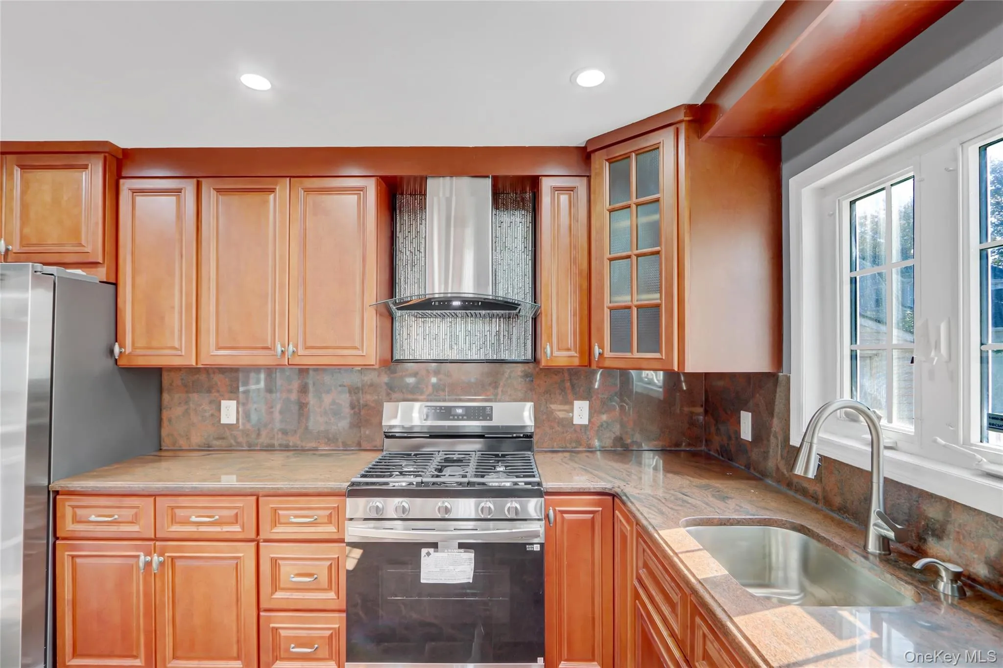 Kitchen featuring recessed lighting, a stainless-steel range with overhead hood, a stainless-steel sink with a gooseneck faucet, light wood-finish cabinetry, and stone-finish countertops and backsplash Kitchen featuring recessed lighting, a stainless-steel range with overhead hood, a stainless-steel sink with a gooseneck faucet, light wood-finish cabinetry, and stone-finish countertops and backsplash