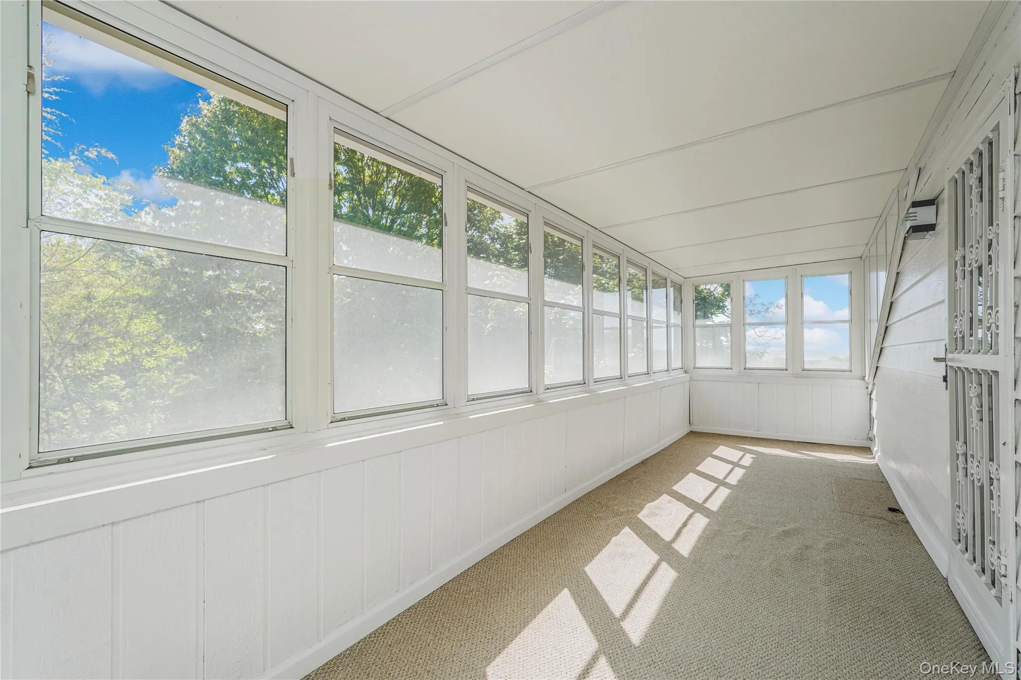 Sunroom featuring extensive window-lined walls, white beadboard wainscoting, white siding, white ceiling panels, and neutral carpet flooring Sunroom featuring extensive window-lined walls, white beadboard wainscoting, white siding, white ceiling panels, and neutral carpet flooring