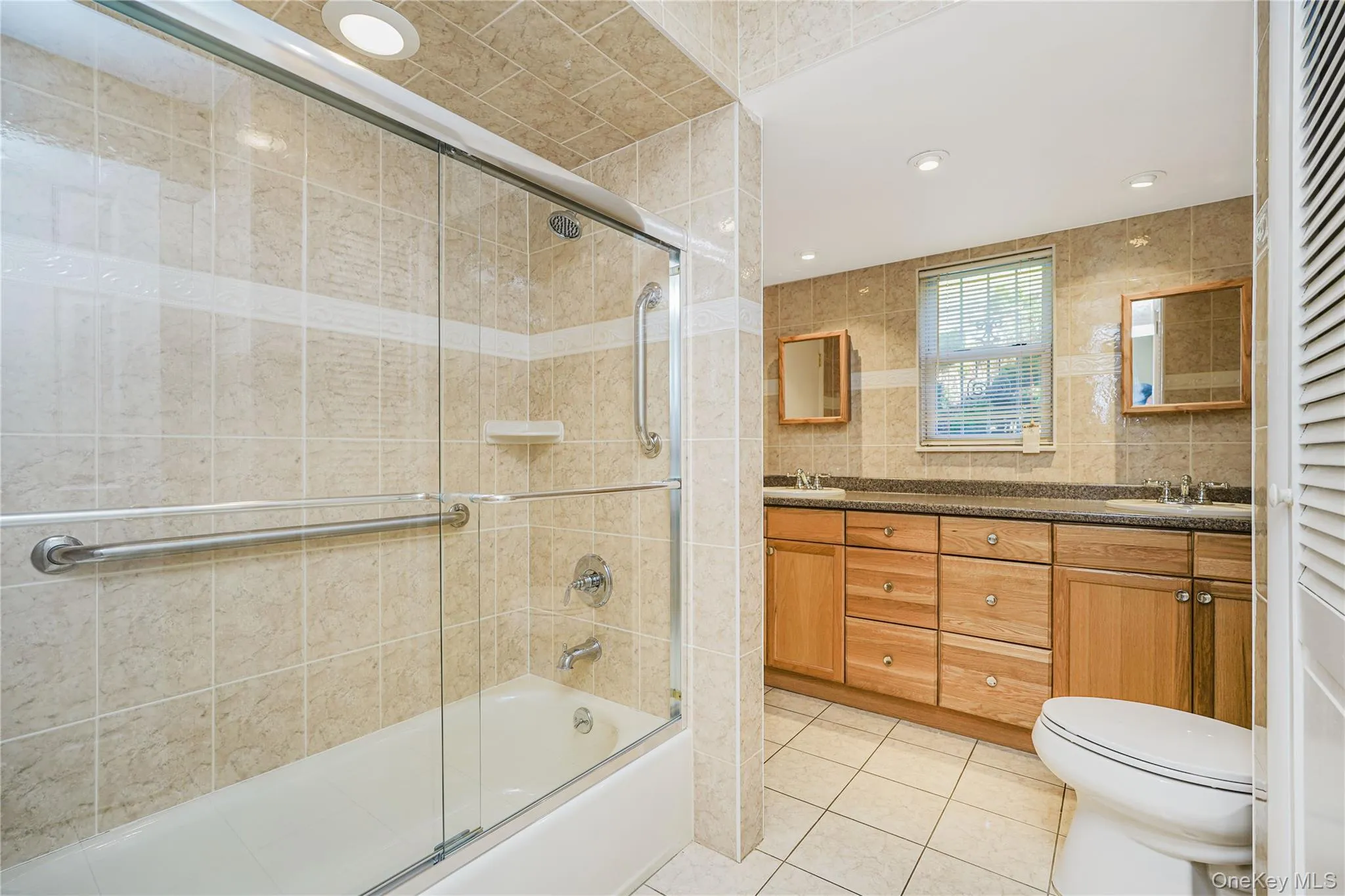 Bathroom featuring a tub/shower combination with glass enclosure, dual vanity with wood-finish cabinetry, and recessed lighting Bathroom featuring a tub/shower combination with glass enclosure, dual vanity with wood-finish cabinetry, and recessed lighting