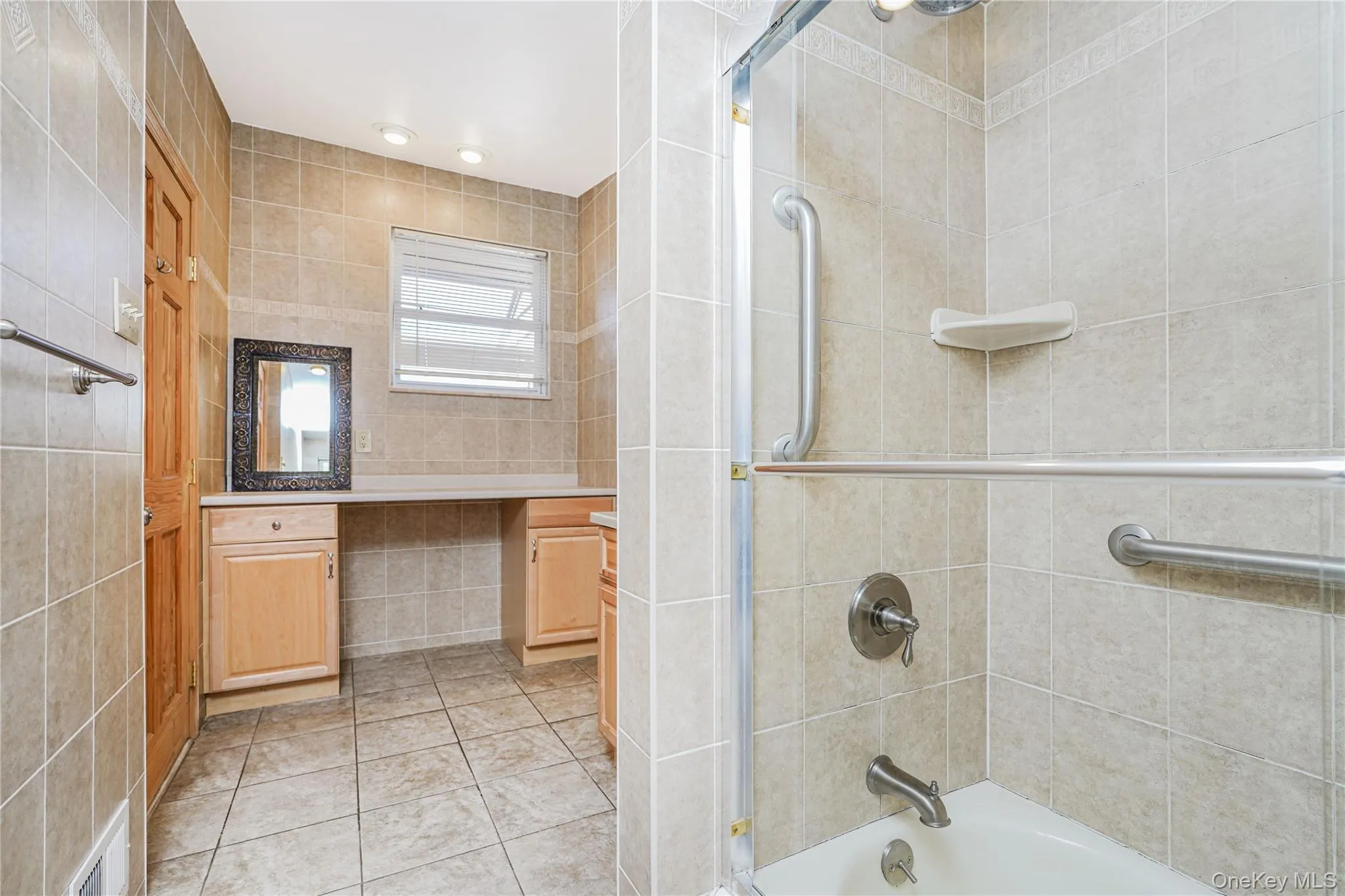 Bathroom featuring neutral-toned floor and wall tiling, a built-in vanity with light wood cabinetry, a window with blinds, and a shower-over-tub with grab bars and a clear enclosure Bathroom featuring neutral-toned floor and wall tiling, a built-in vanity with light wood cabinetry, a window with blinds, and a shower-over-tub with grab bars and a clear enclosure