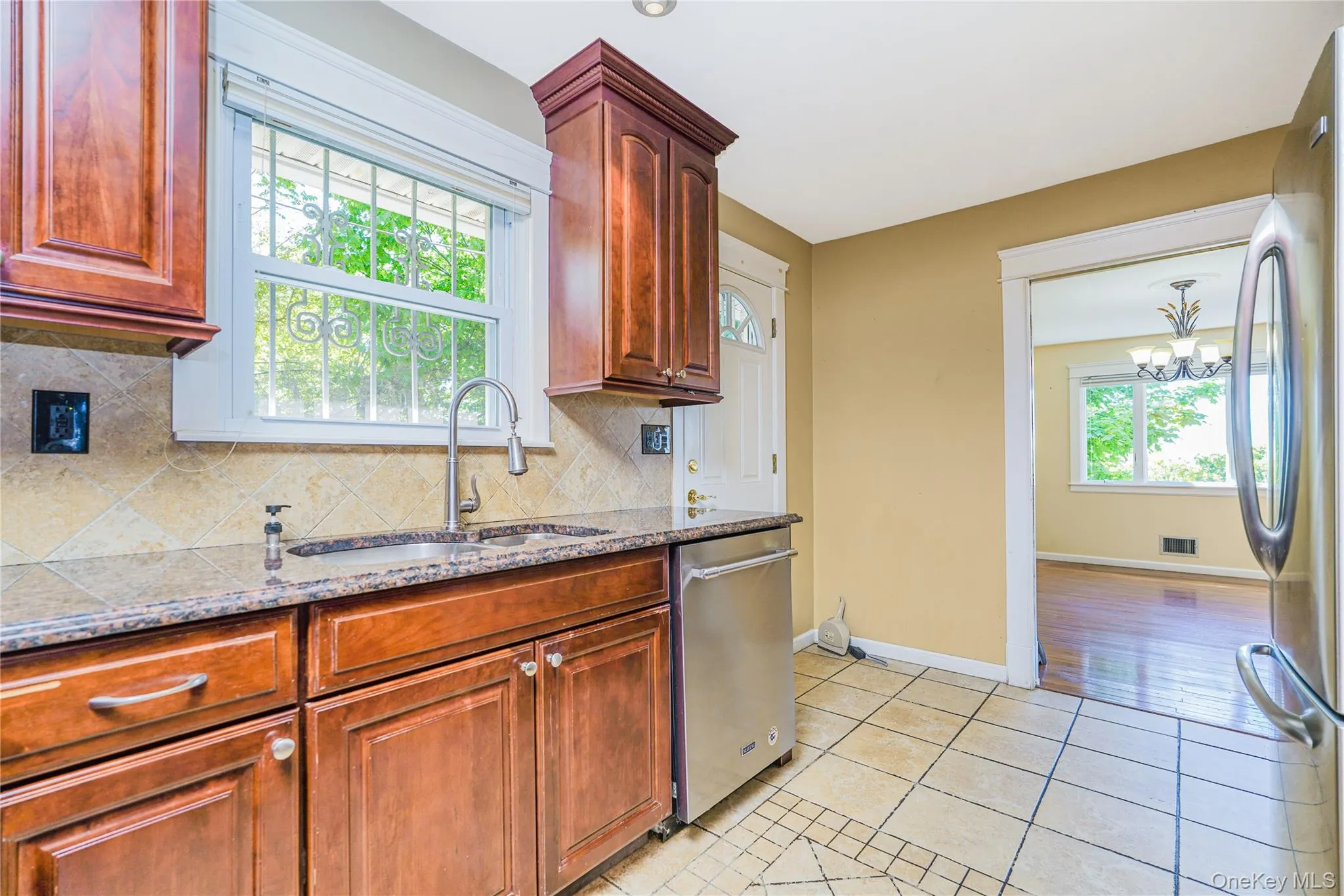 Kitchen featuring wood cabinetry, granite countertops, tile backsplash, stainless steel sink with a pull-down faucet, and tile flooring Kitchen featuring wood cabinetry, granite countertops, tile backsplash, stainless steel sink with a pull-down faucet, and tile flooring
