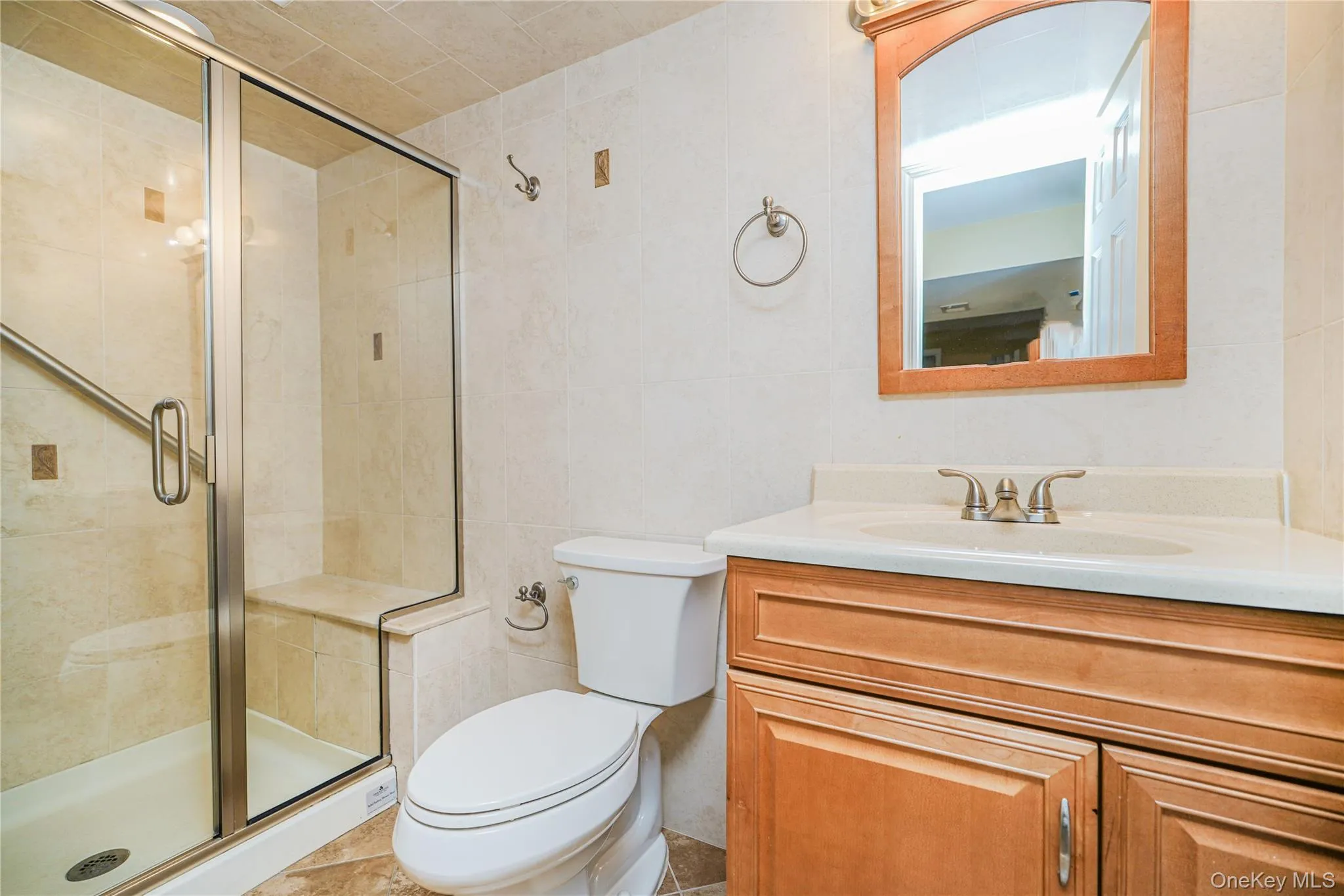 Bathroom featuring a glass-enclosed shower with a built-in bench, a toilet, a vanity with a white countertop and integrated sink, a wood-framed mirror, and tiled flooring Bathroom featuring a glass-enclosed shower with a built-in bench, a toilet, a vanity with a white countertop and integrated sink, a wood-framed mirror, and tiled flooring