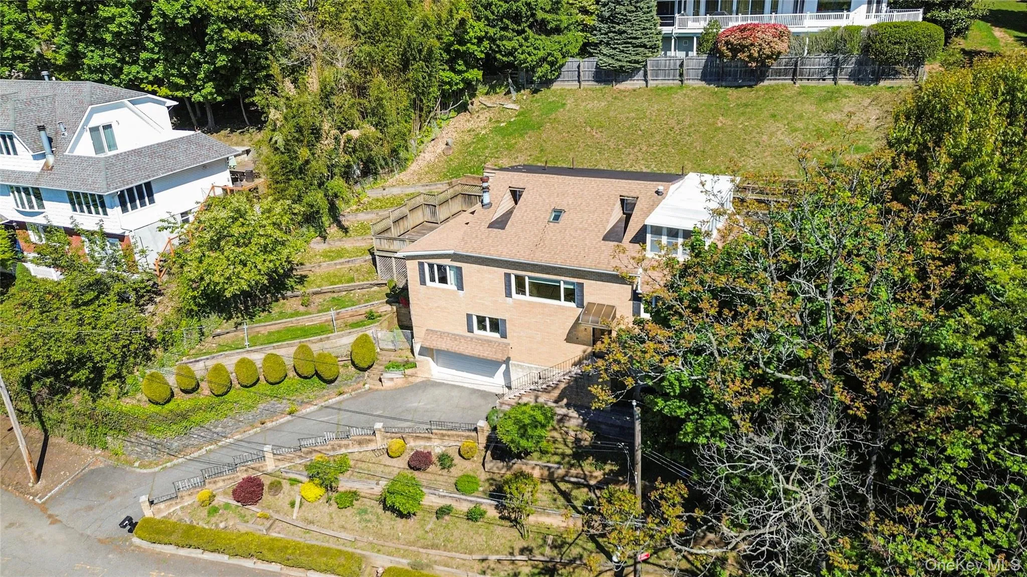 Brick facade residence featuring a multi-level terraced landscape, an attached garage, dormer windows, and a paved driveway Brick facade residence featuring a multi-level terraced landscape, an attached garage, dormer windows, and a paved driveway
