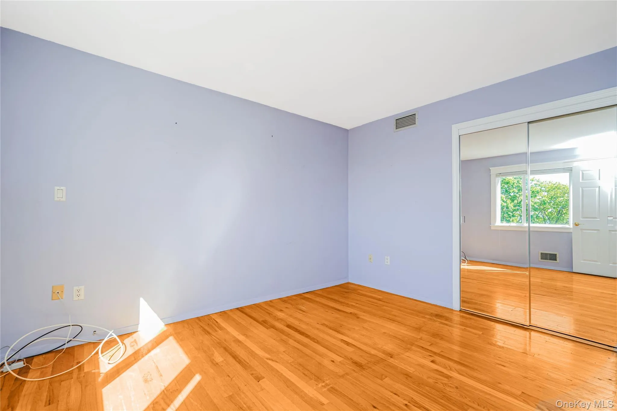 Interior room featuring wood-finish flooring, a mirrored sliding closet door, and a window with tree scenery Interior room featuring wood-finish flooring, a mirrored sliding closet door, and a window with tree scenery