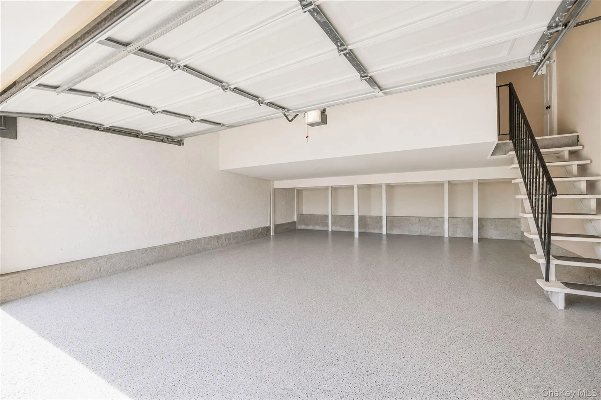 Spacious garage featuring a light gray speckled epoxy floor finish, an overhead door with exposed track, integrated wall shelving, and a white staircase with a black metal railing Spacious garage featuring a light gray speckled epoxy floor finish, an overhead door with exposed track, integrated wall shelving, and a white staircase with a black metal railing