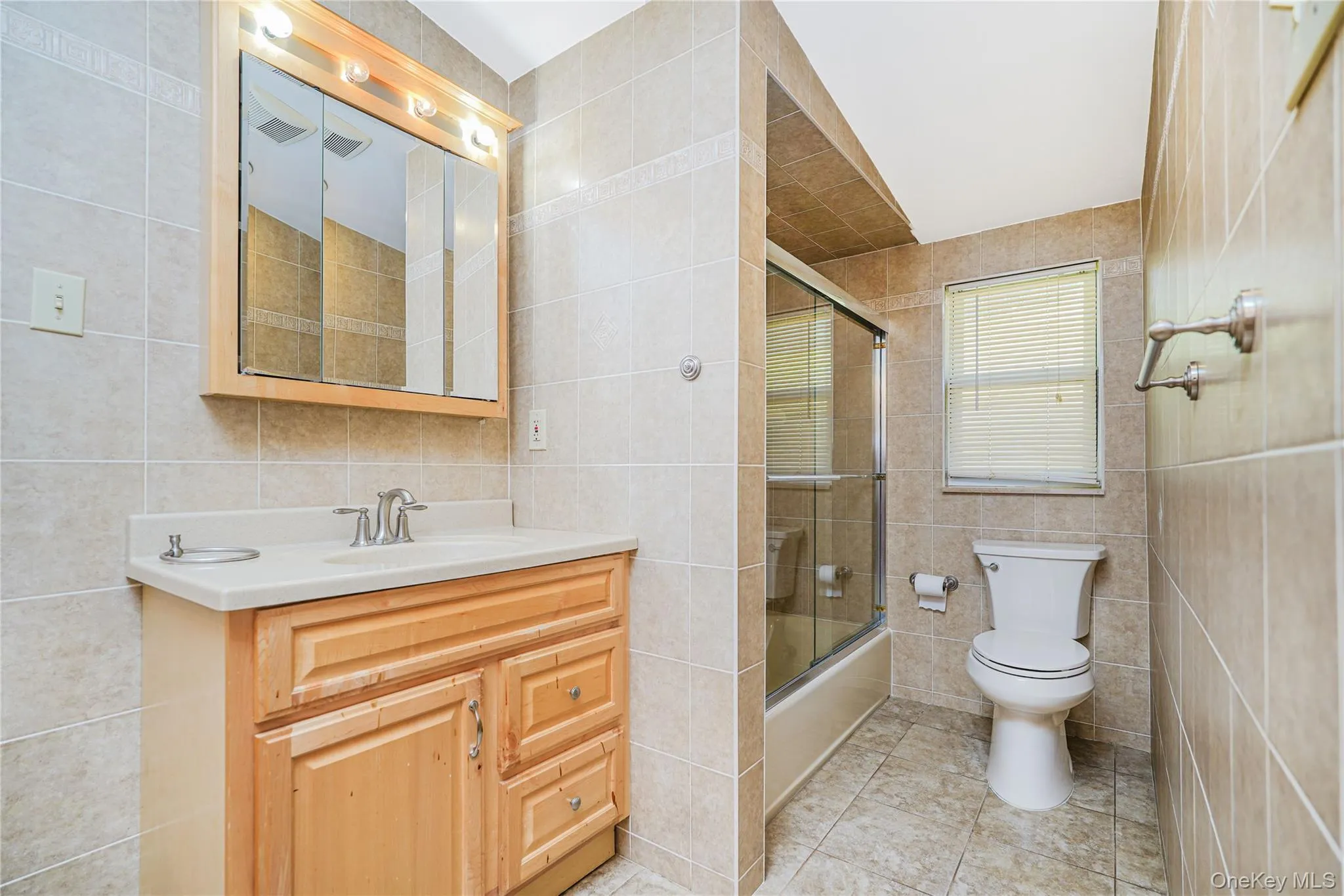 Bathroom featuring a light wood-finish vanity with an integrated sink, a mirrored medicine cabinet with overhead lighting, a tiled shower-tub combination with sliding glass doors, a window with blinds, and floor-to-ceiling tilework Bathroom featuring a light wood-finish vanity with an integrated sink, a mirrored medicine cabinet with overhead lighting, a tiled shower-tub combination with sliding glass doors, a window with blinds, and floor-to-ceiling tilework