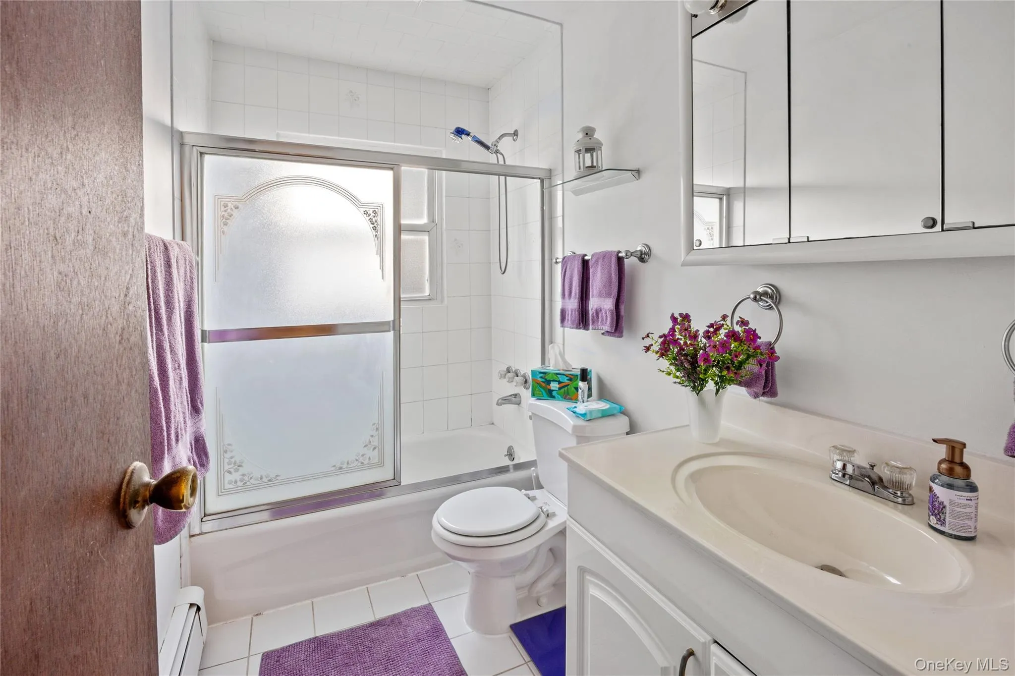 White tiled bathroom featuring a built-in bathtub with sliding glass doors, a white vanity with sink, and a mirrored medicine cabinet White tiled bathroom featuring a built-in bathtub with sliding glass doors, a white vanity with sink, and a mirrored medicine cabinet