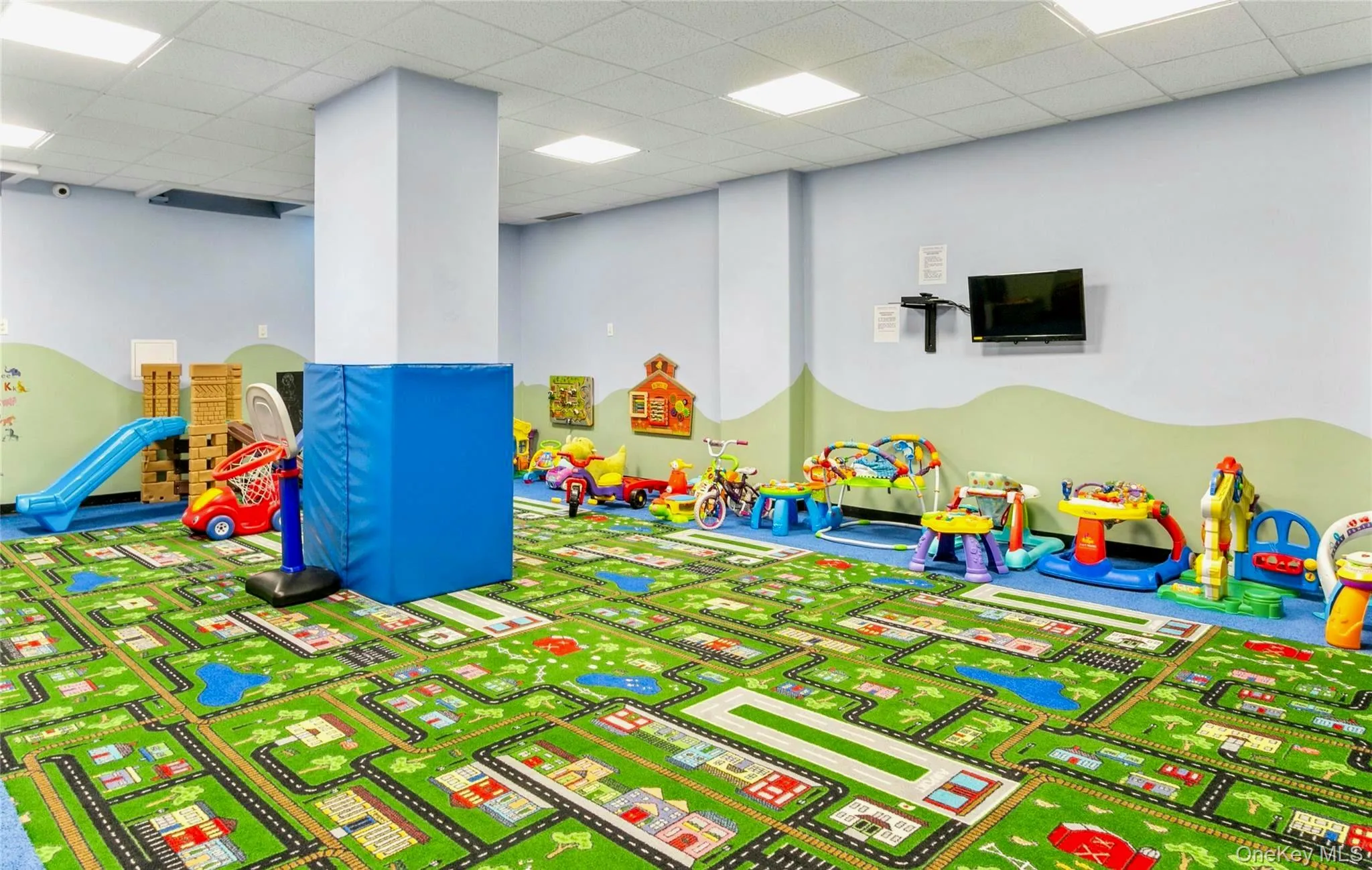 Recreational room featuring a durable patterned carpet, light blue walls with a green accent mural, recessed ceiling lighting, and a wall-mounted television Recreational room featuring a durable patterned carpet, light blue walls with a green accent mural, recessed ceiling lighting, and a wall-mounted television