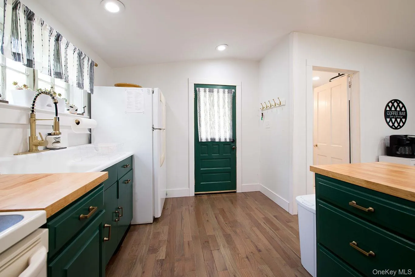 Kitchen featuring wood-finish flooring, dark green cabinetry with brass hardware, light-toned butcher block countertops, and a white sink with a gold-tone faucet Kitchen featuring wood-finish flooring, dark green cabinetry with brass hardware, light-toned butcher block countertops, and a white sink with a gold-tone faucet