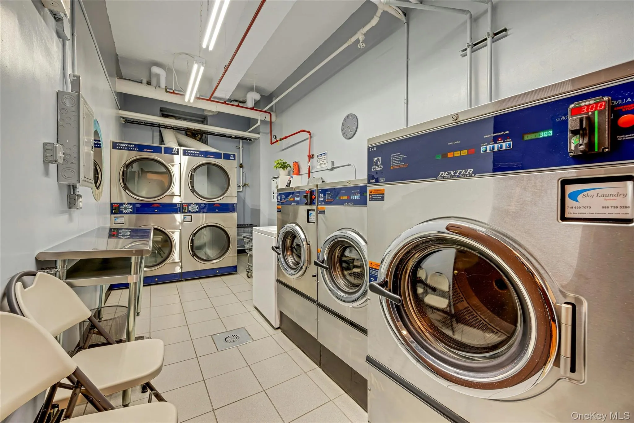 On-site laundry facility featuring multiple commercial-grade washers and dryers, tiled flooring, and a stainless steel folding table On-site laundry facility featuring multiple commercial-grade washers and dryers, tiled flooring, and a stainless steel folding table